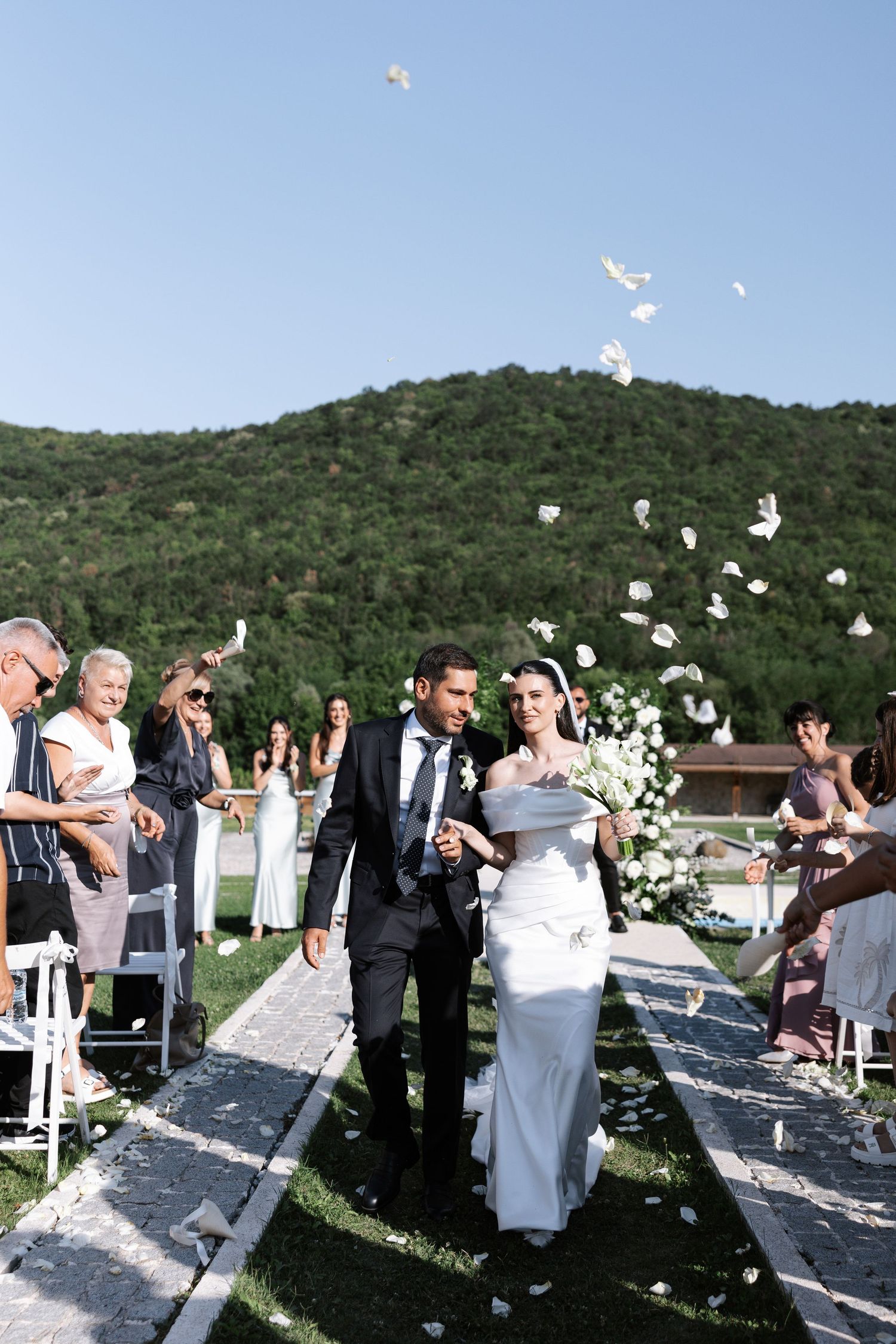 Newlyweds walk down an outdoor aisle while white flower petals float through the air against a mountain backdrop.