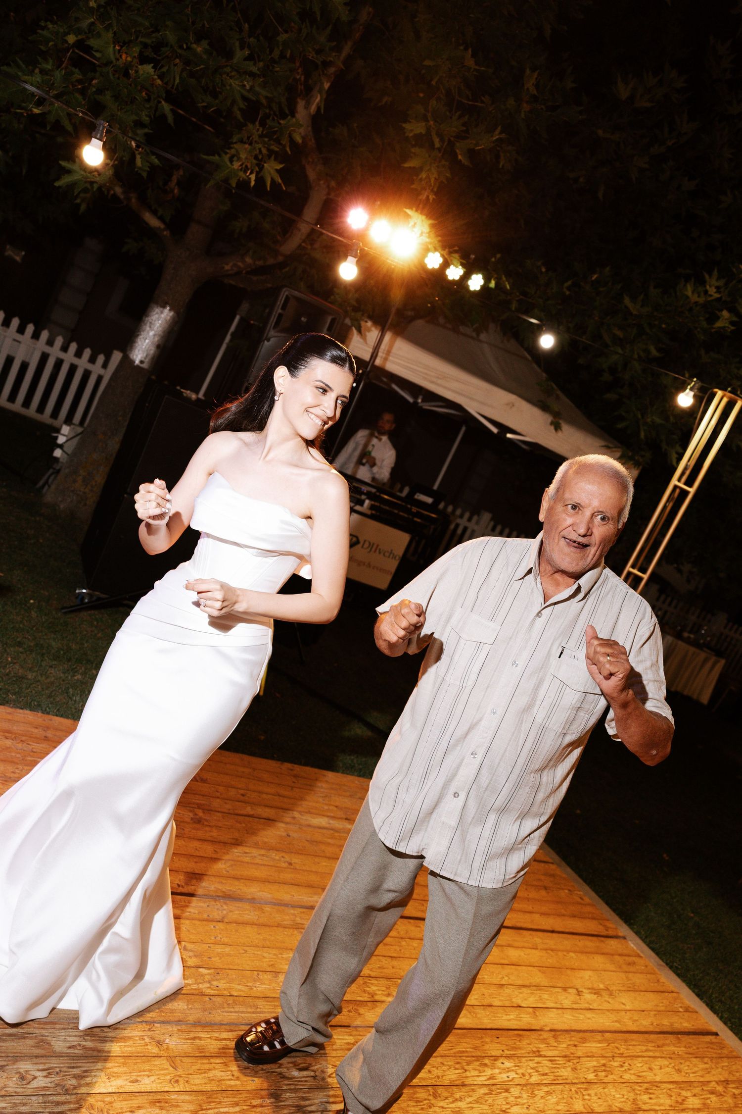 Two people dancing together on a wooden dance floor at an outdoor evening reception with string lights overhead.