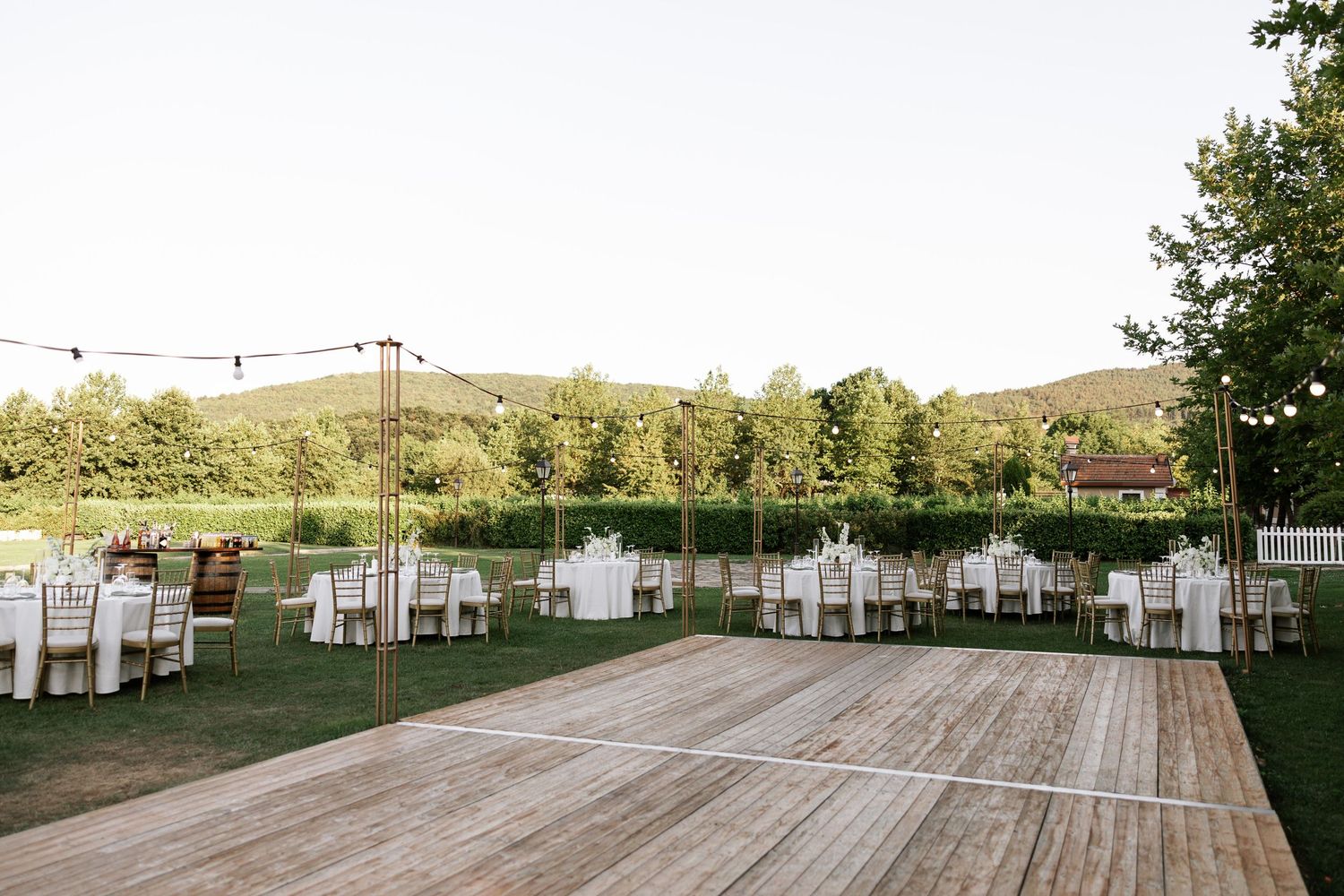 Outdoor wedding venue with wooden dance floor and white chairs set up on grass under string lights at dusk.