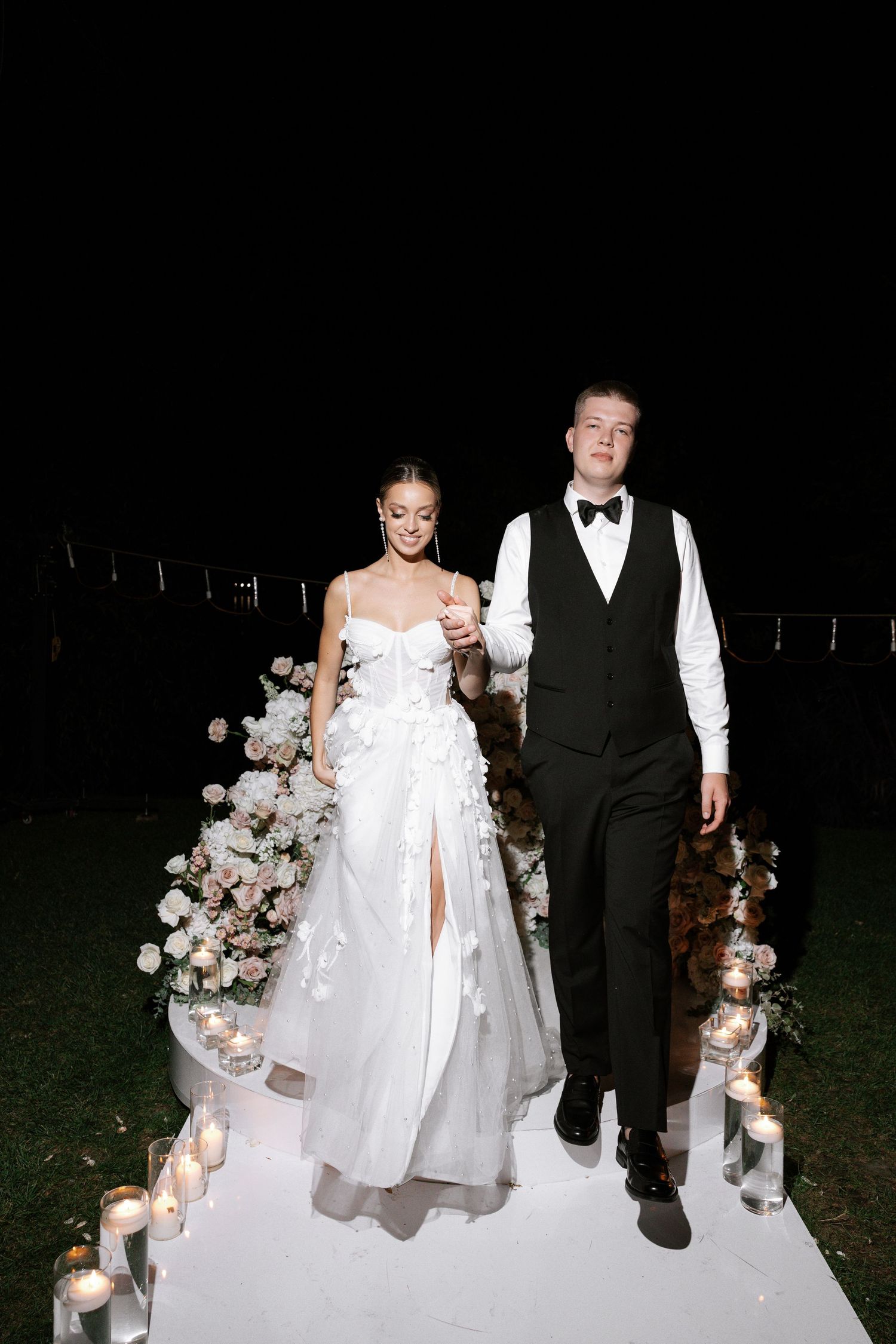 Evening wedding portrait of couple on illuminated white walkway with floral decorations.