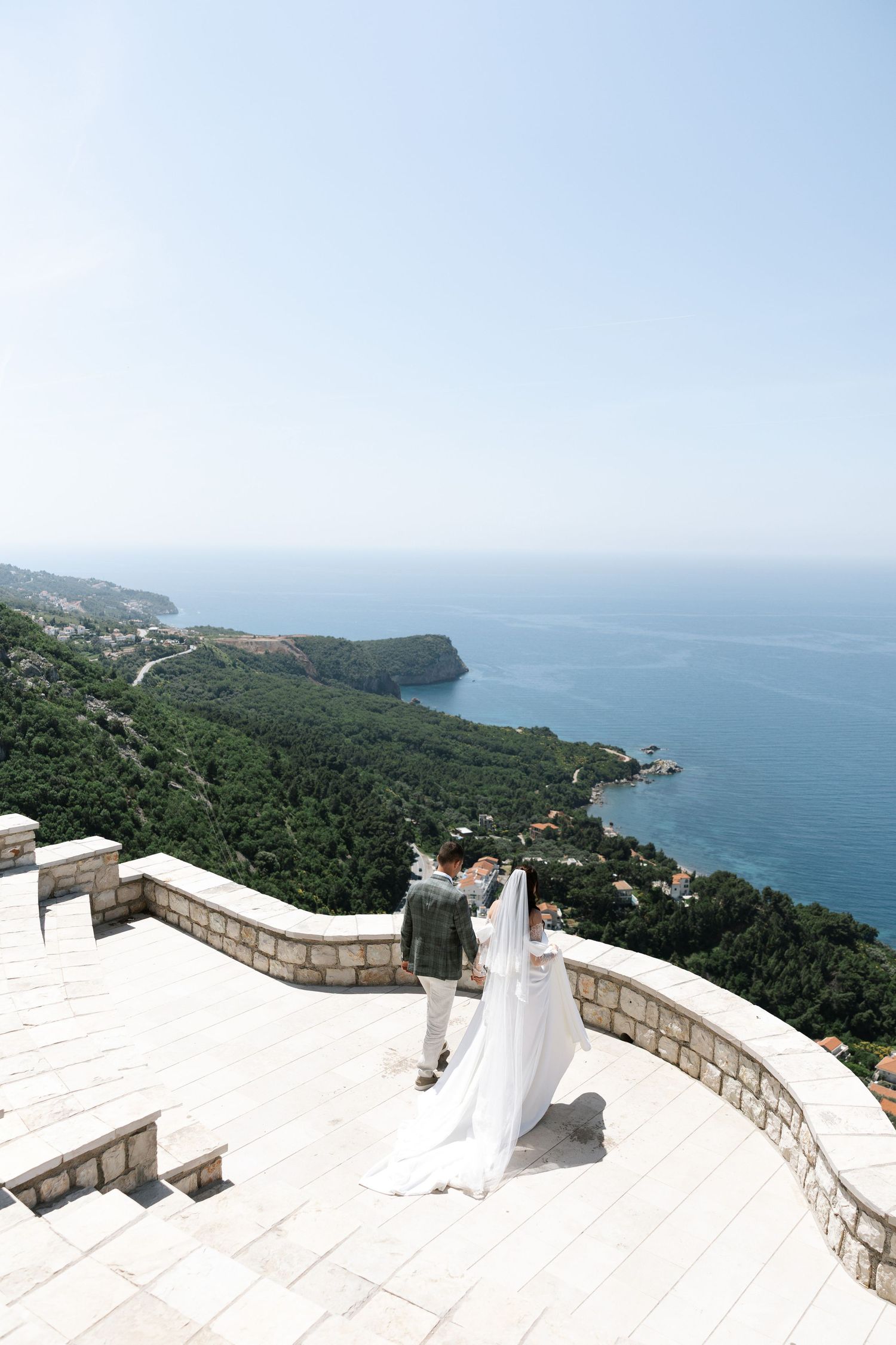 Panoramic coastal view from a white stone terrace overlooking blue ocean waters and forested cliffs.