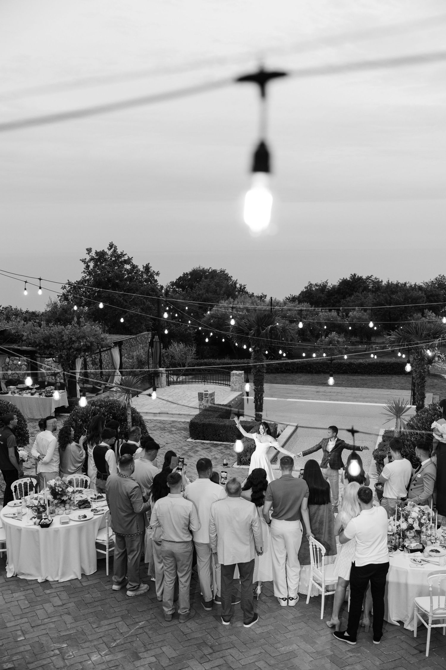 Wedding guests gather on an outdoor patio at dusk under string lights near dining tables at a reception venue.