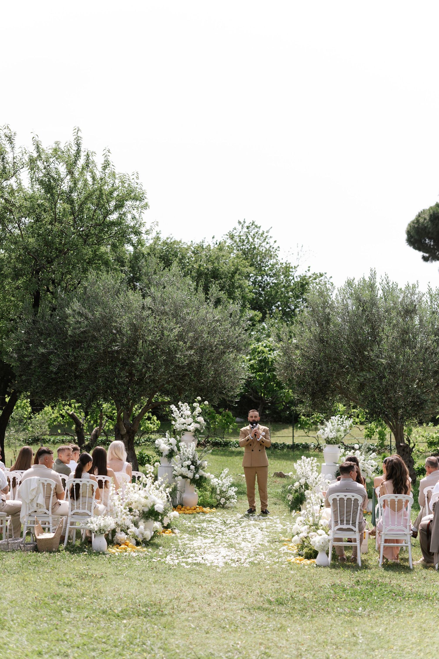 An intimate outdoor wedding ceremony setup in an orchard with white chairs and scattered flower petals on the grass.