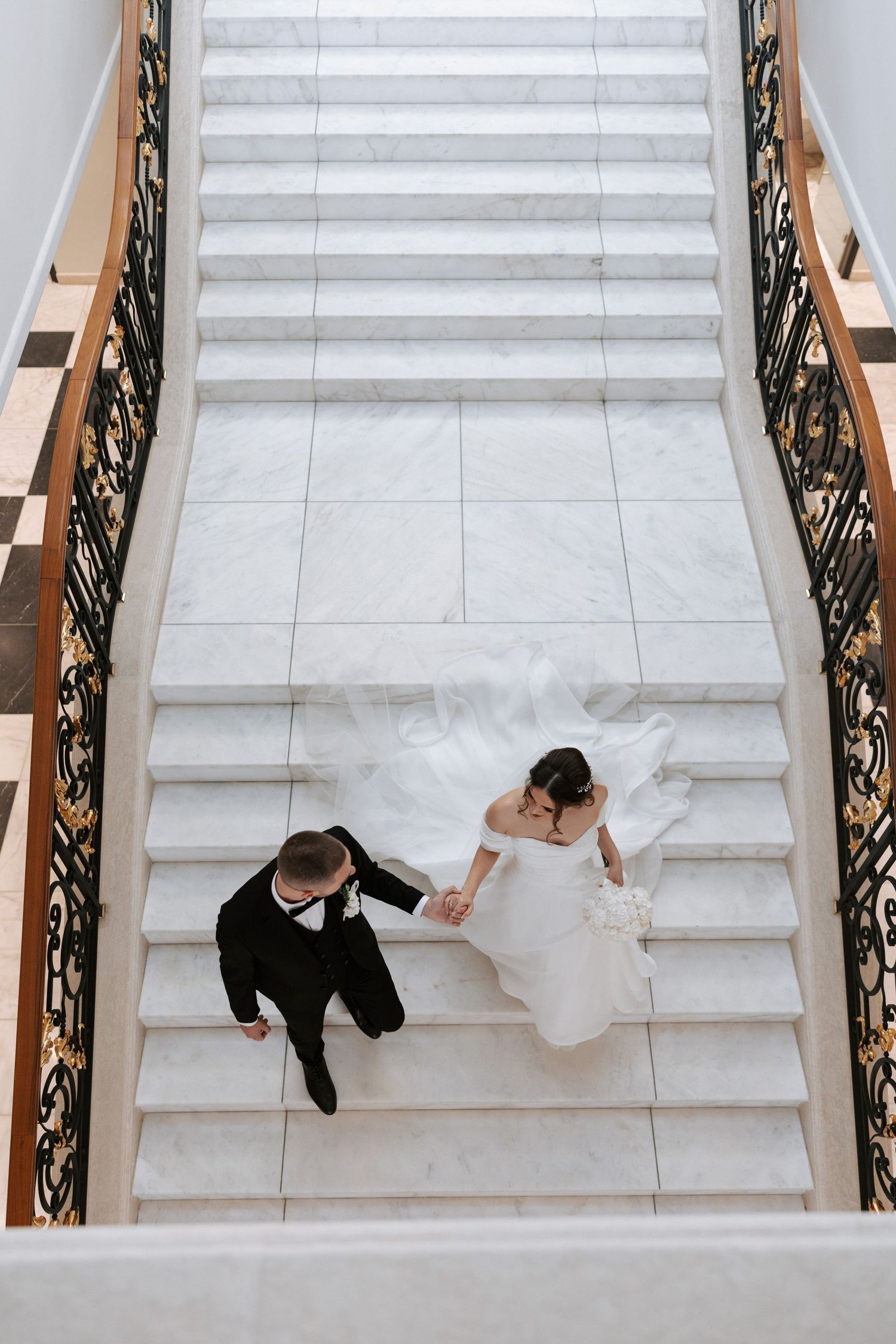 Bird's eye view of couple descending elegant marble staircase with ornate iron railings.