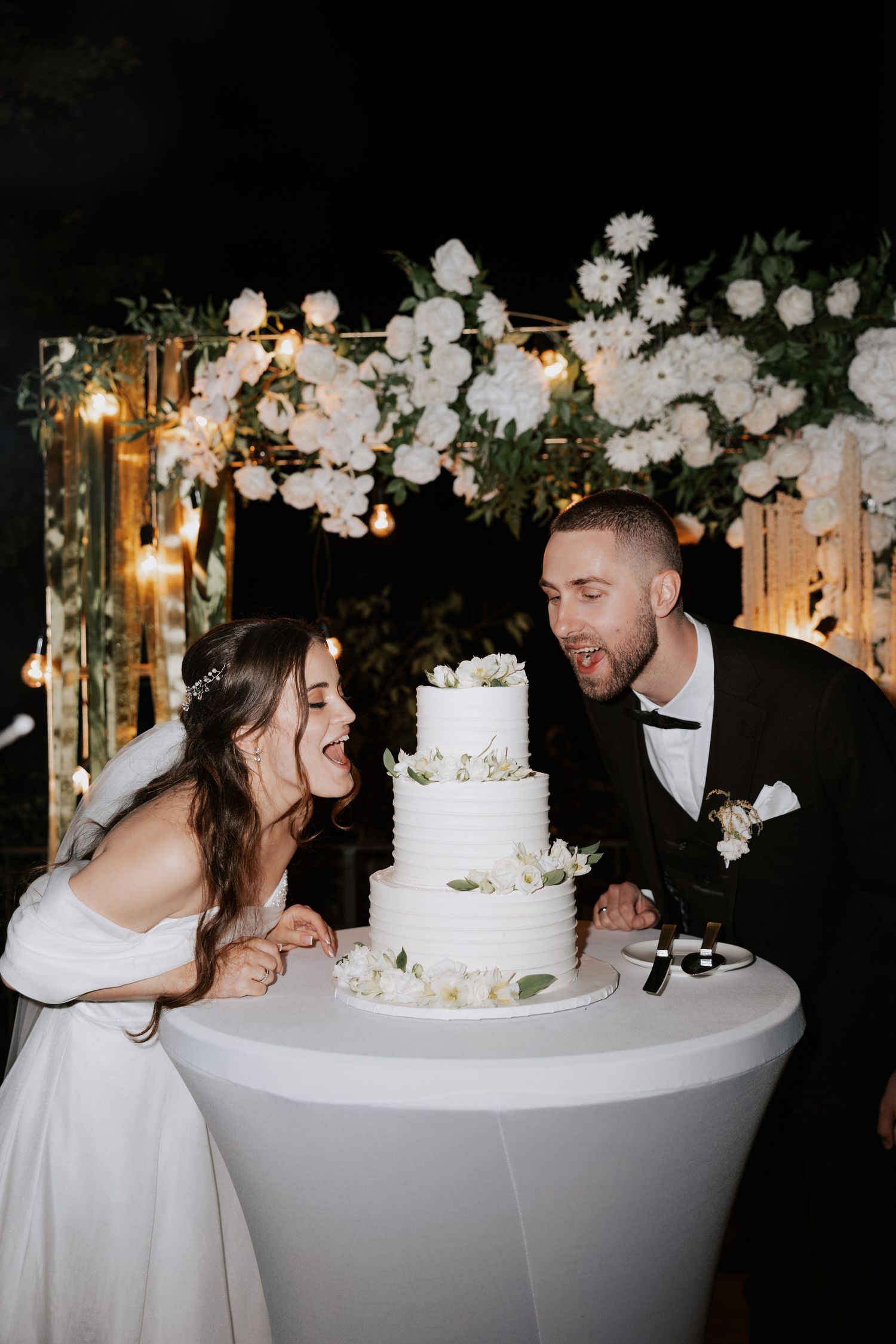 A couple shares a joyful moment while cutting their elegant white wedding cake under floral decorations.