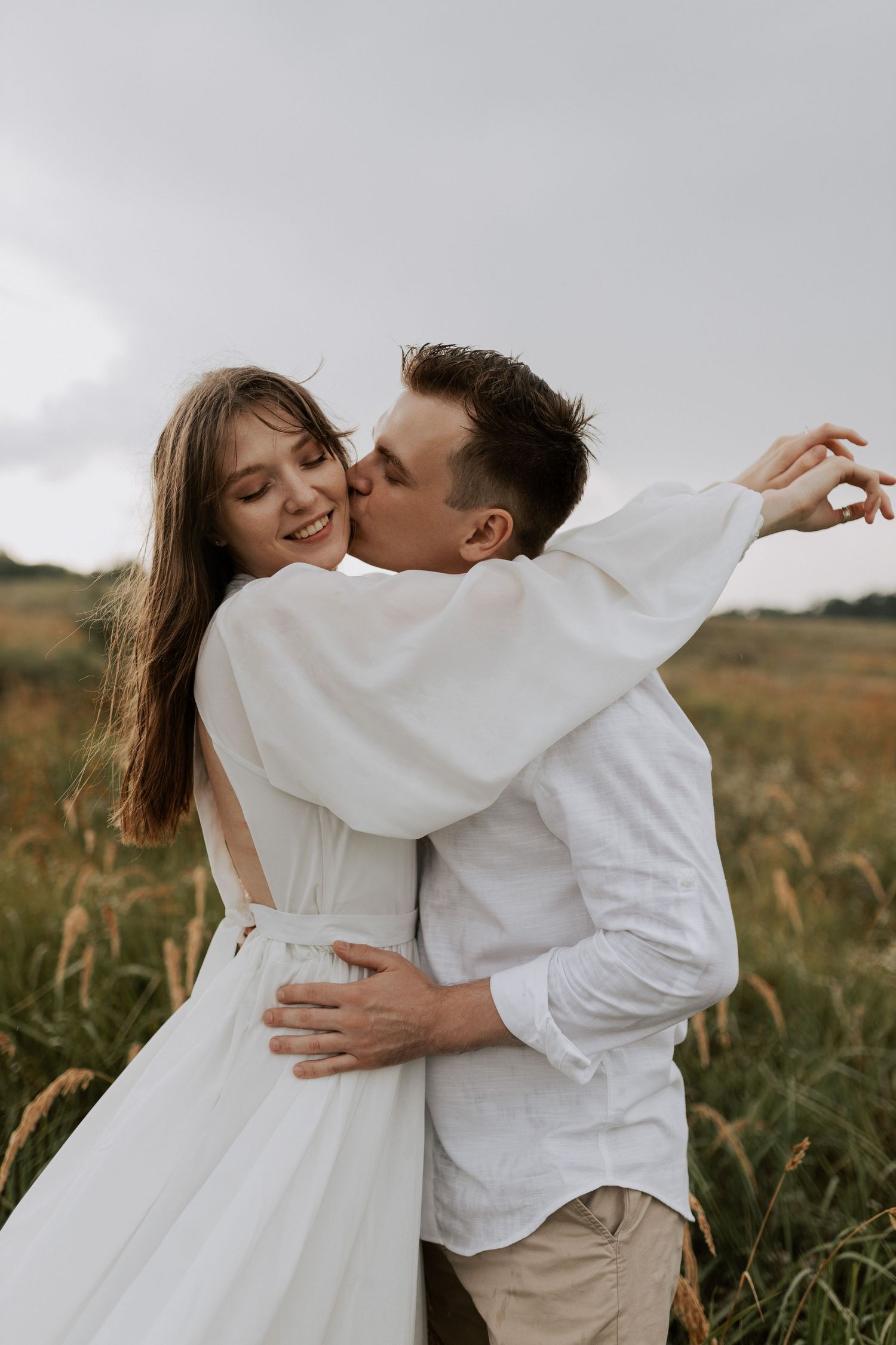 Couple sharing intimate moment in field wearing white clothing during outdoor photoshoot.