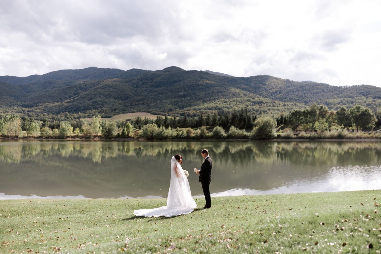A distant view of a wedding couple standing by a peaceful mountain lake.