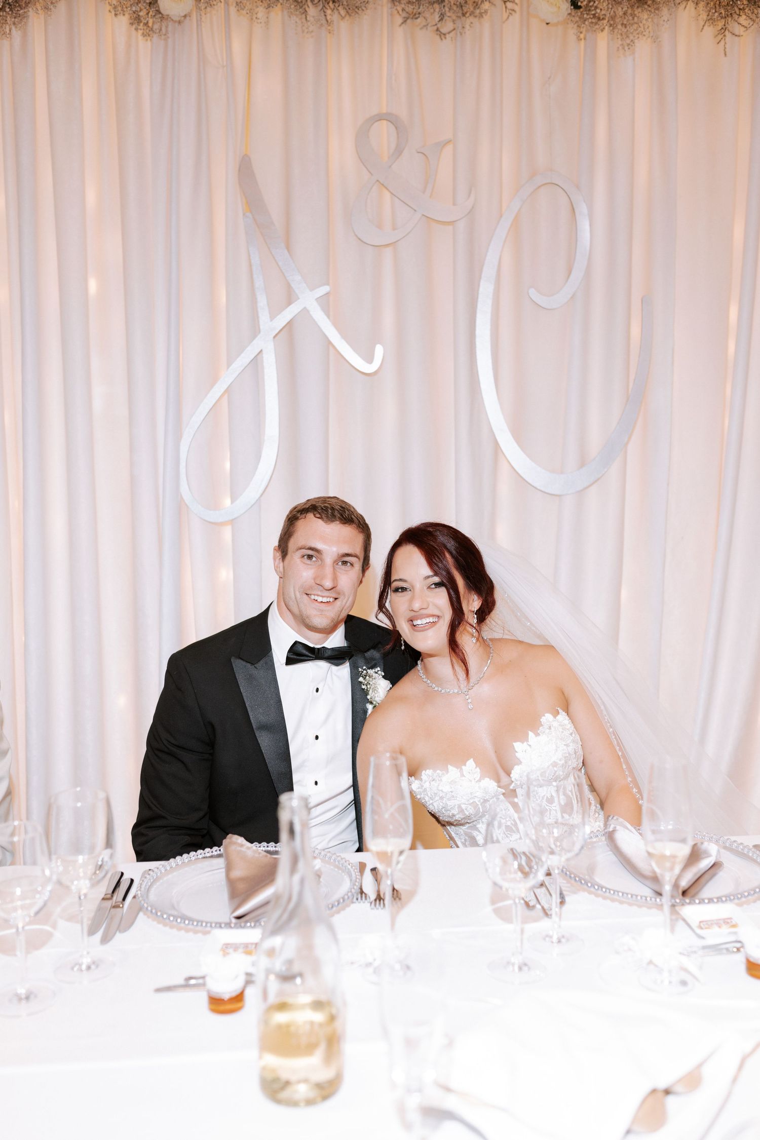 Newlyweds seated at their reception table with elegant white backdrop and monogram details.