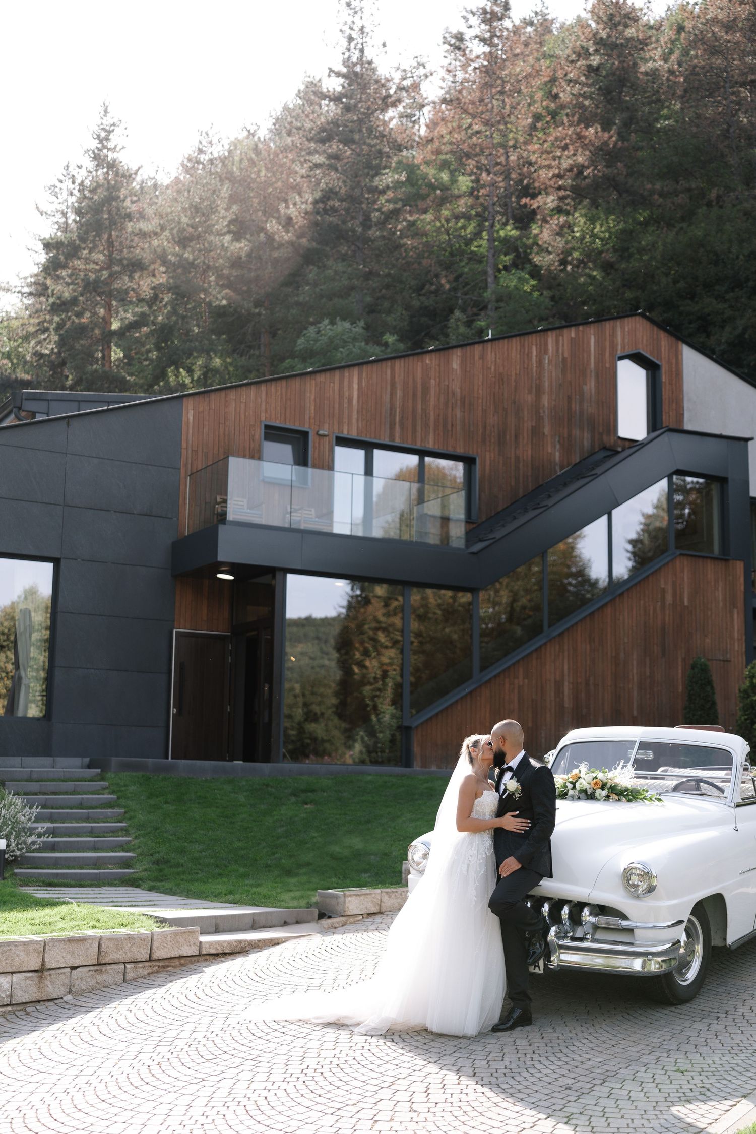 Modern house exterior with dark panels and wood siding features classic white vintage car parked in driveway.