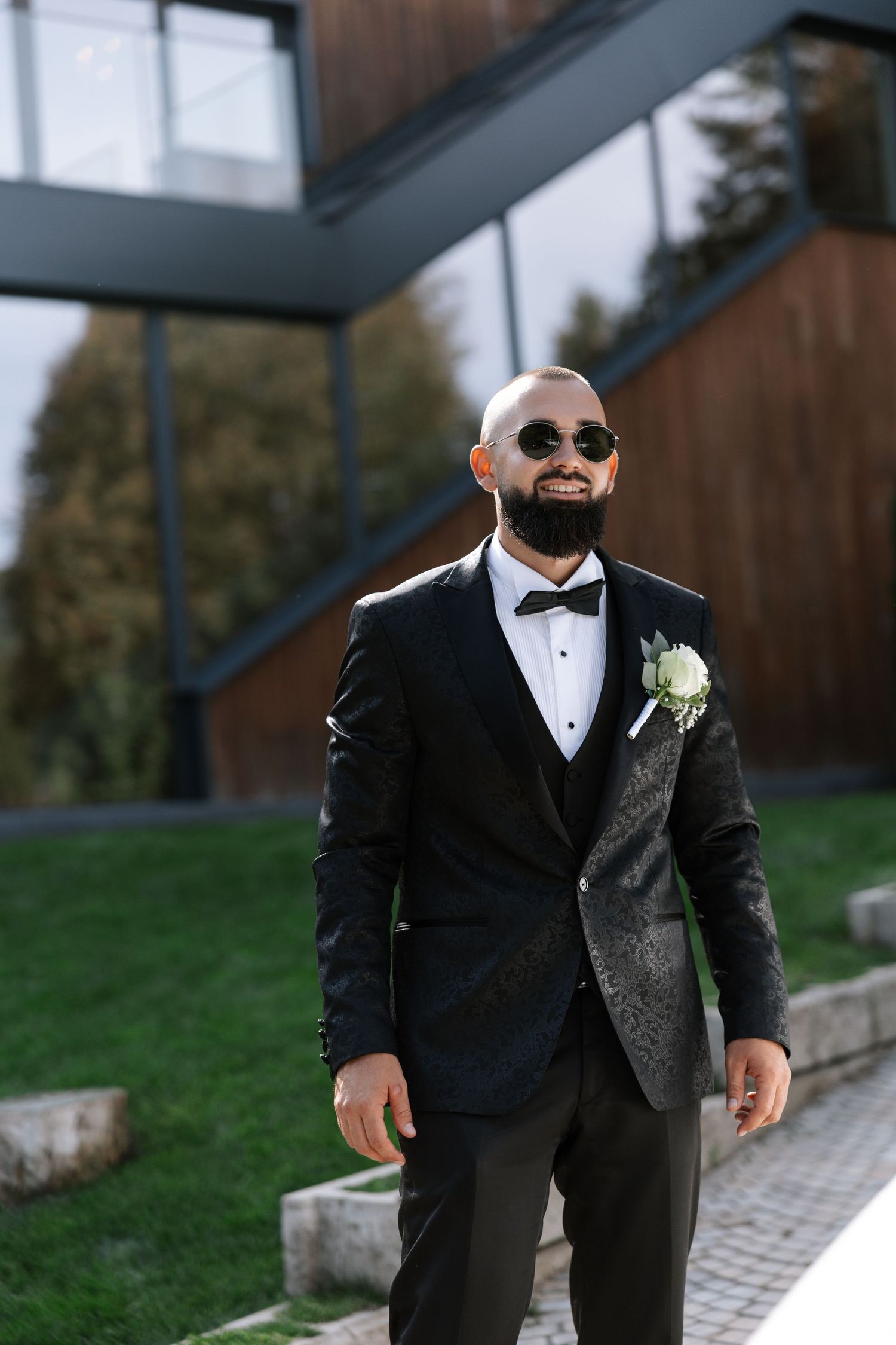 A man in a formal black tuxedo and sunglasses walks along a stone path during a wedding celebration.