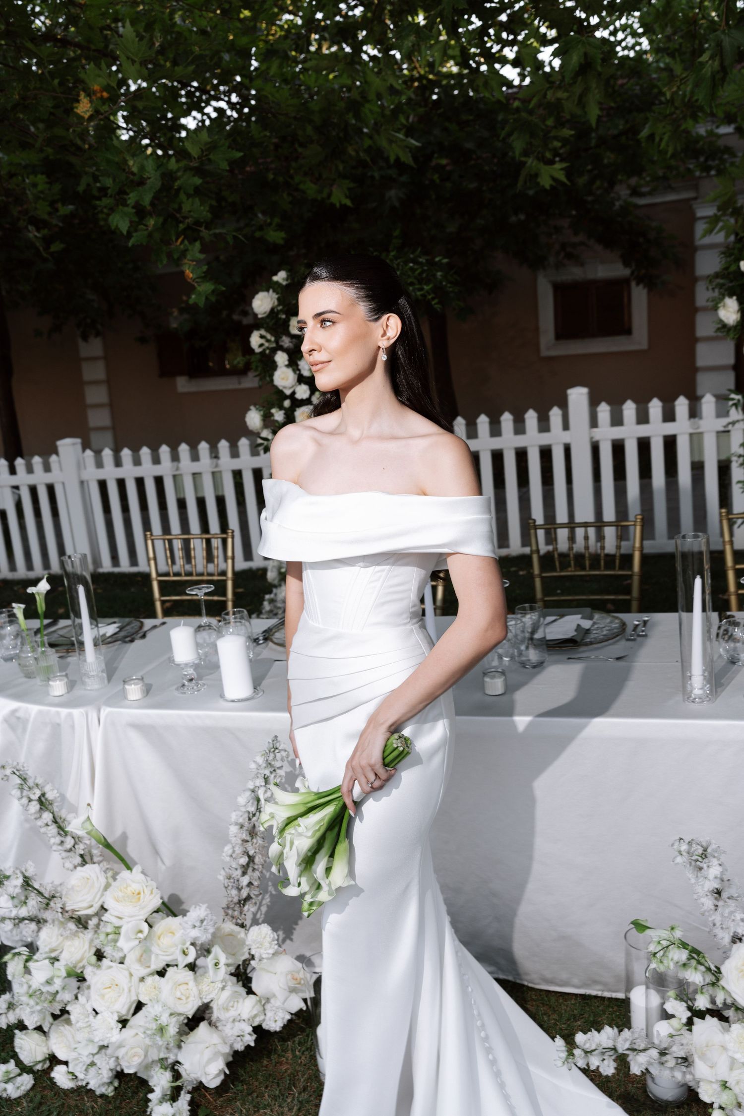 A bride in a white off-shoulder wedding gown poses elegantly against a white picket fence surrounded by floral decorations.