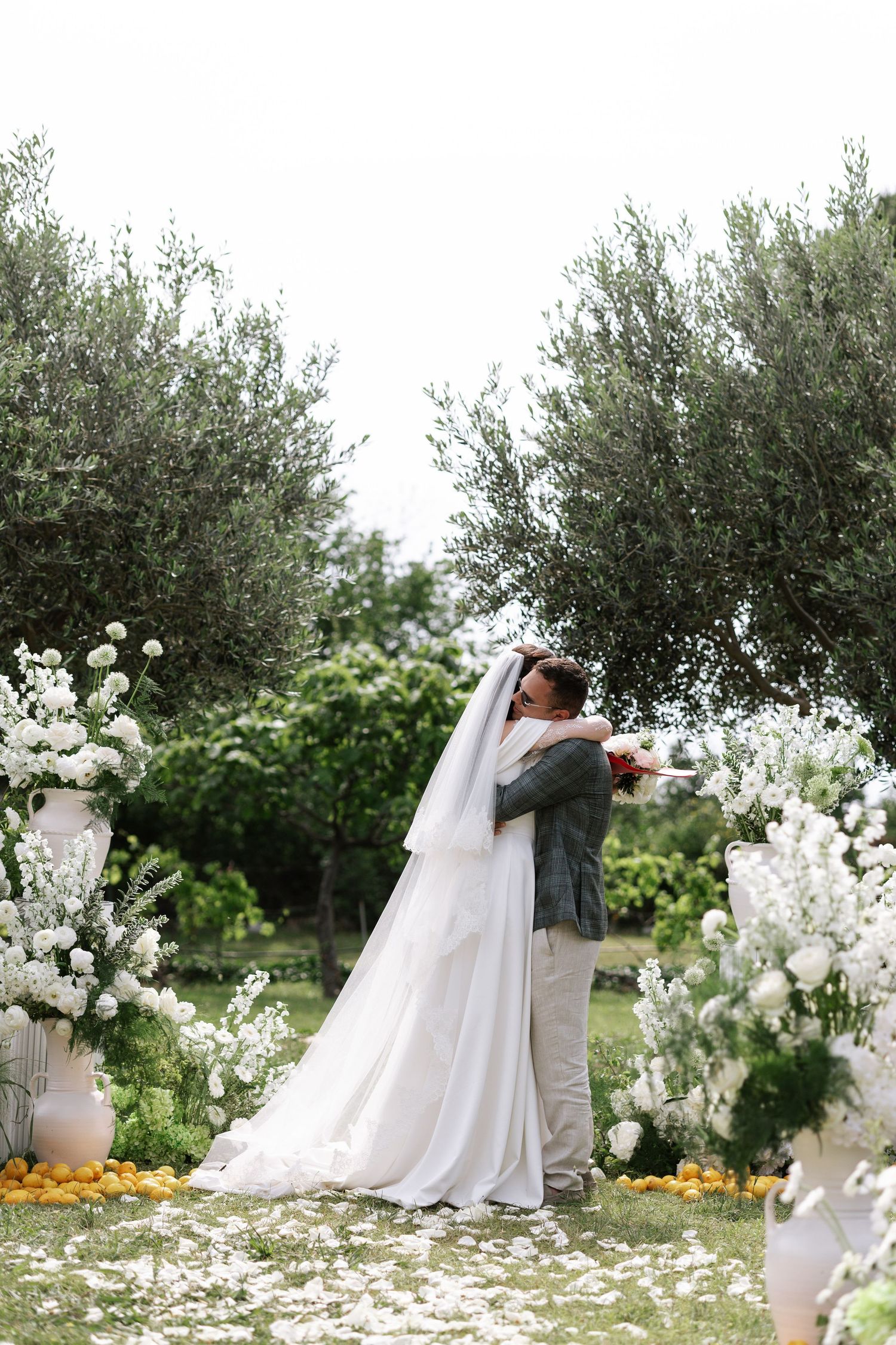A romantic wedding ceremony takes place under an olive tree with white floral arrangements and marigolds in the garden.