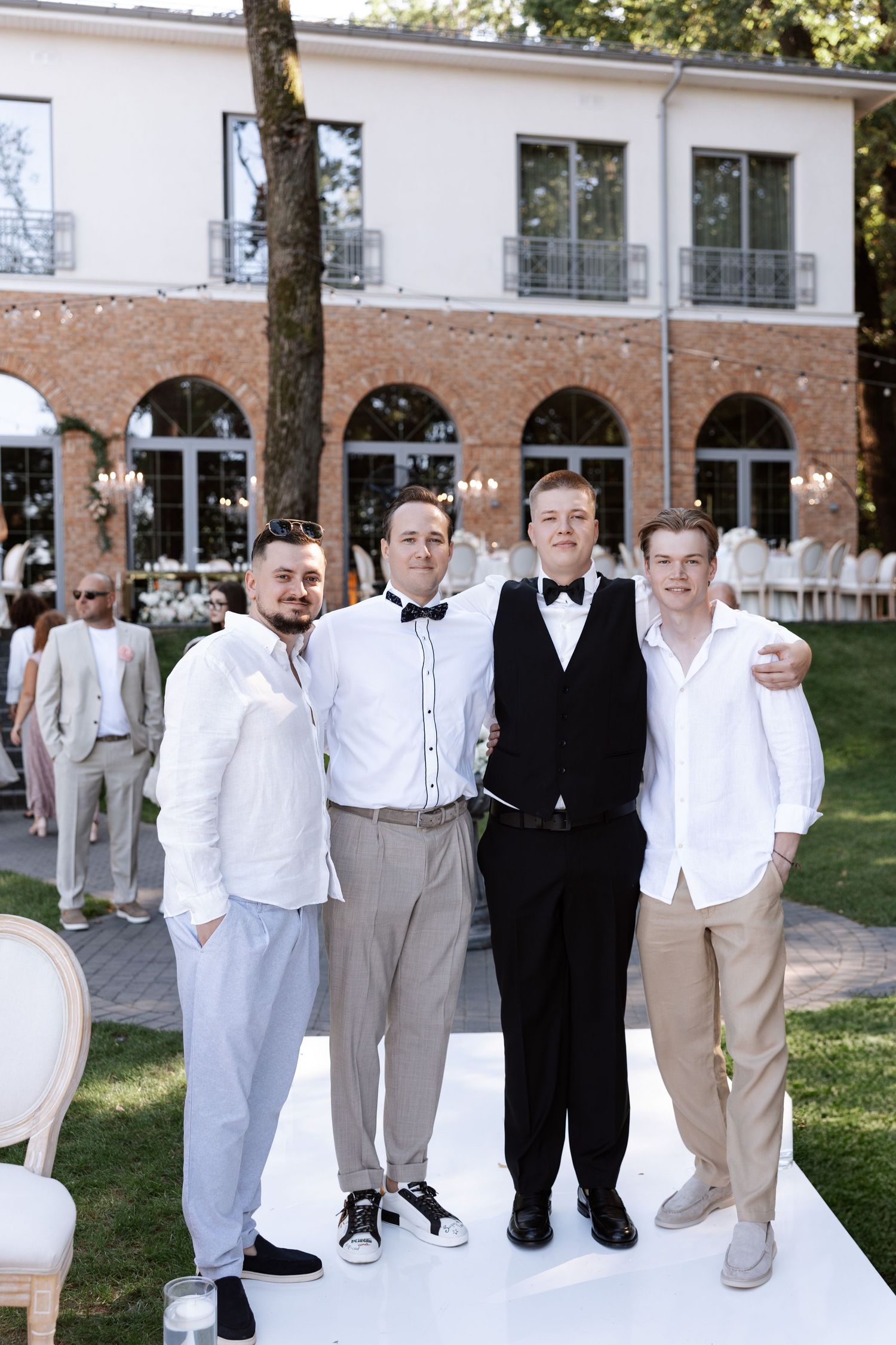 Wedding party groomsmen in formal attire posing together in front of brick venue with arched windows.