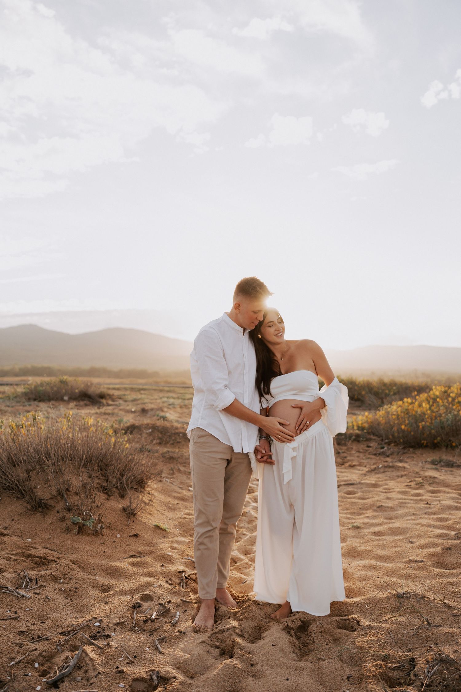 Couple embraces tenderly in desert setting with mountains and sunset behind them.