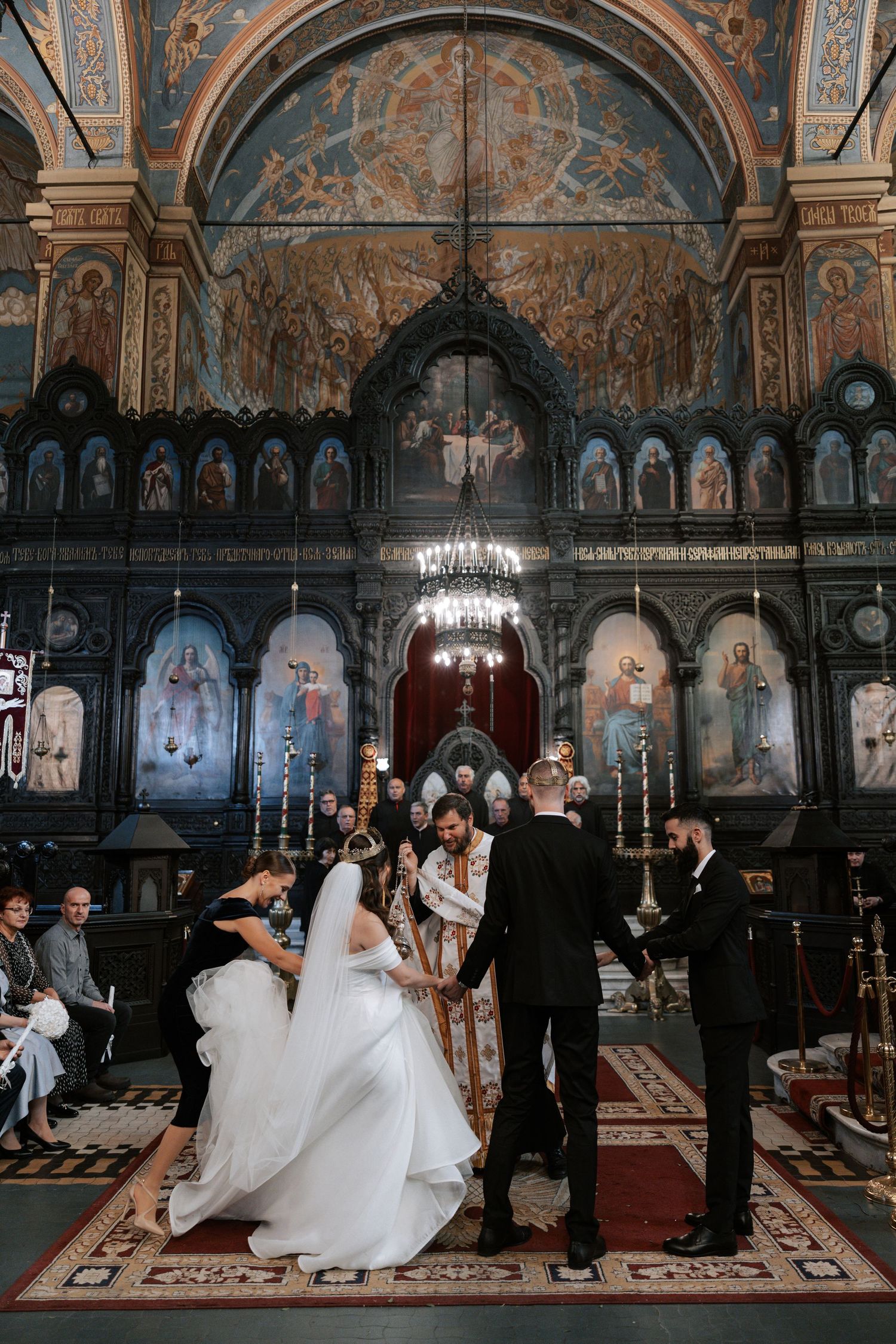 Wedding ceremony in progress at an Orthodox church with elaborate iconostasis and frescoed ceiling.