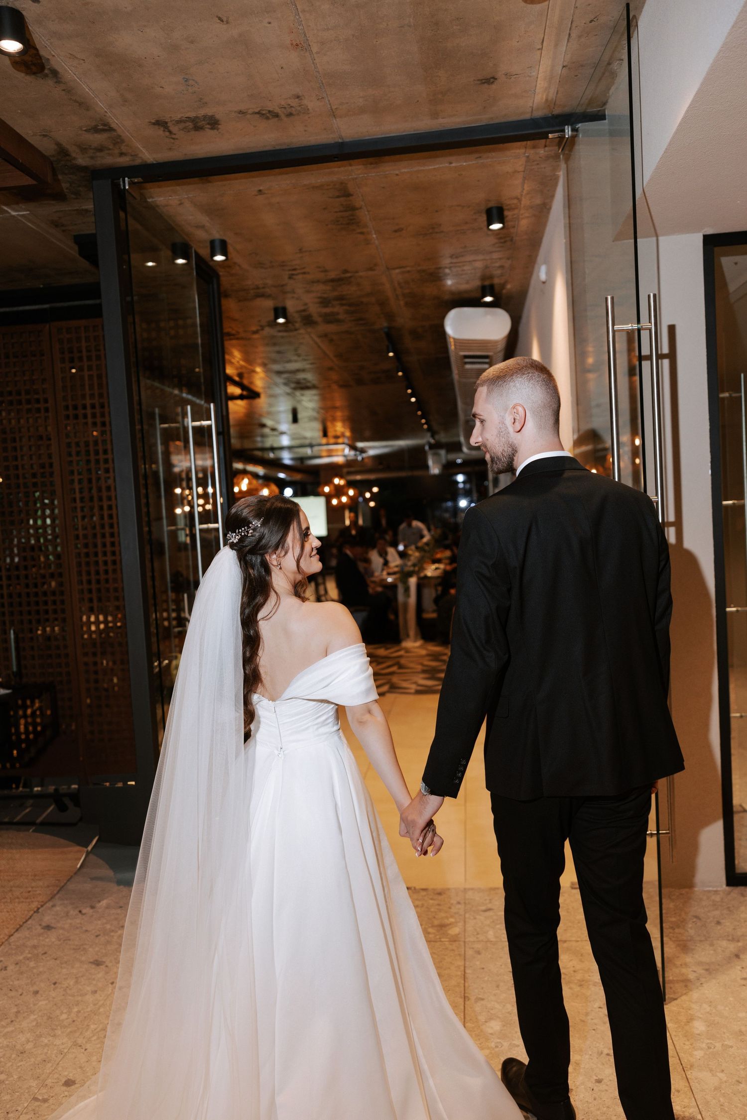 Elegant newlyweds walking hand in hand through a modern venue with dramatic lighting and industrial decor.