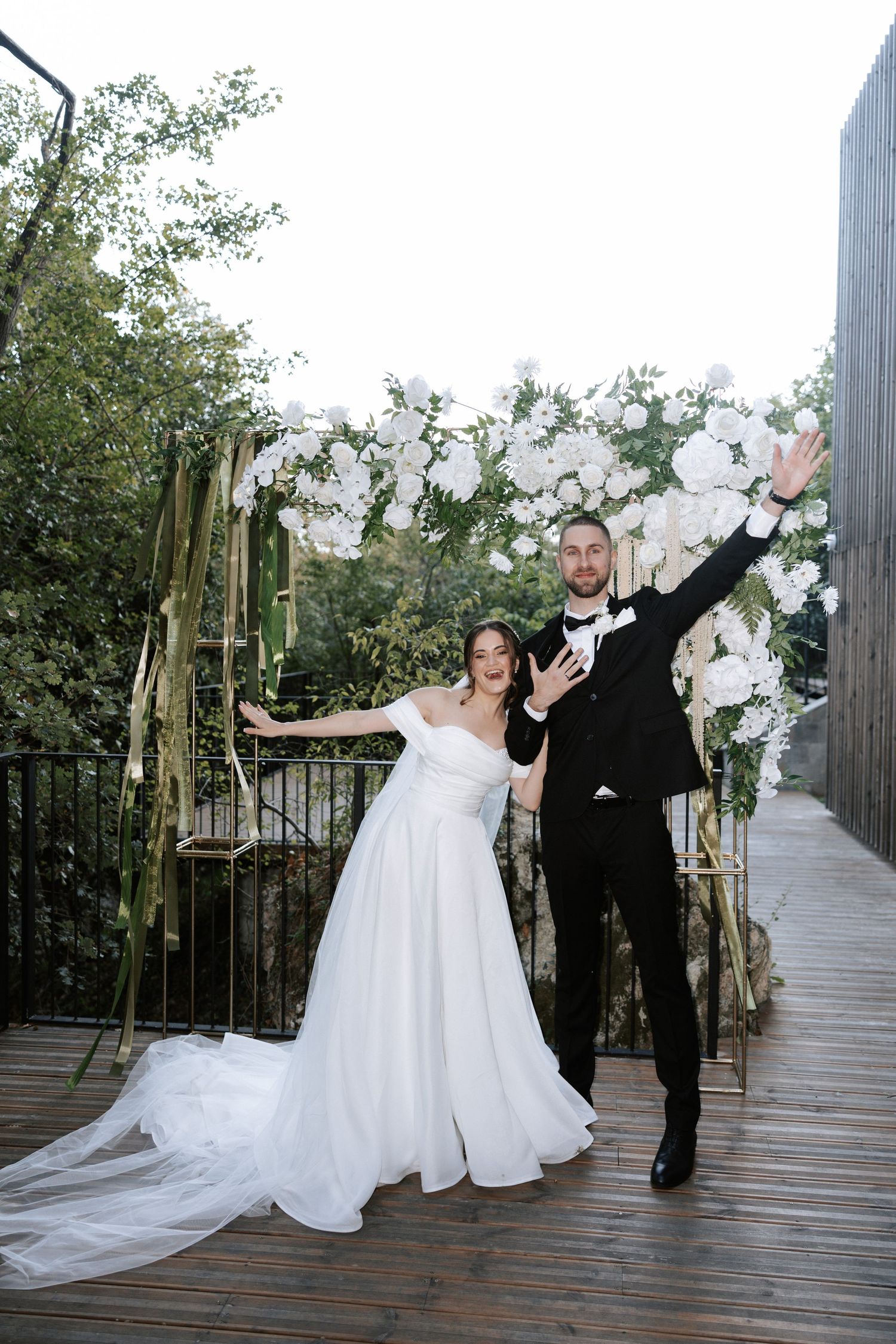 Newlyweds celebrate with joyful poses under a decorative white floral arch.
