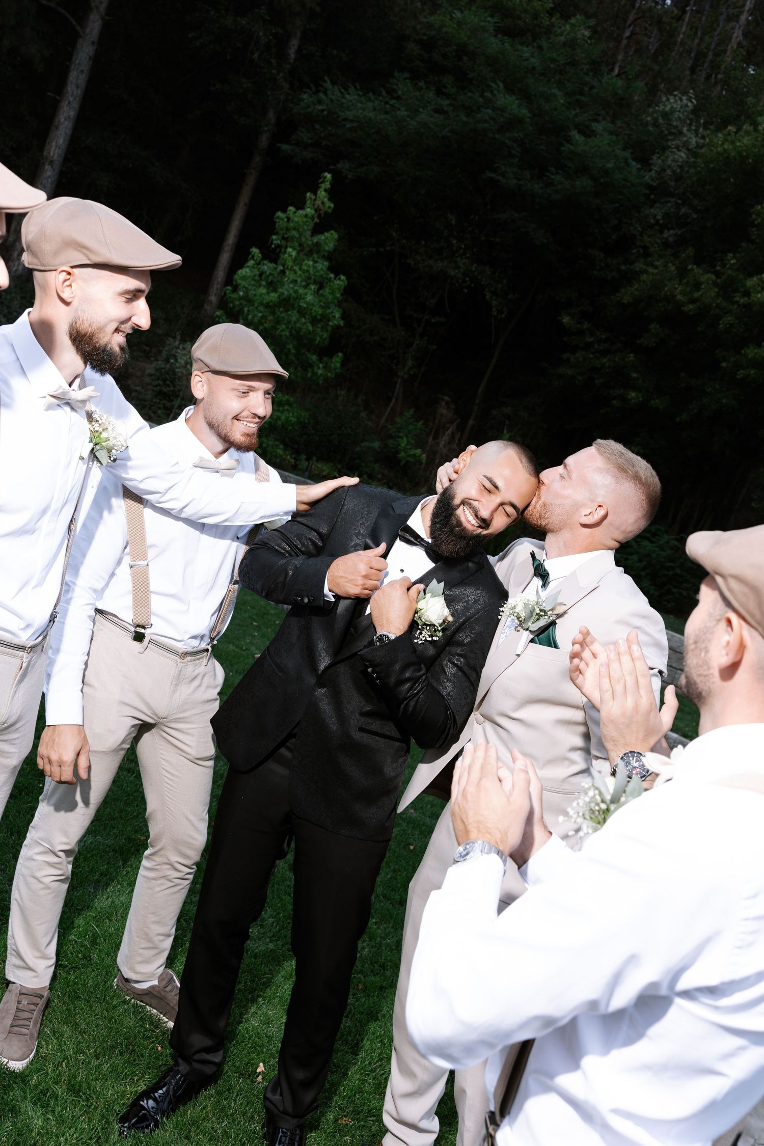 A group of groomsmen in light colored suits and dark suits pose playfully for photos in an outdoor setting.