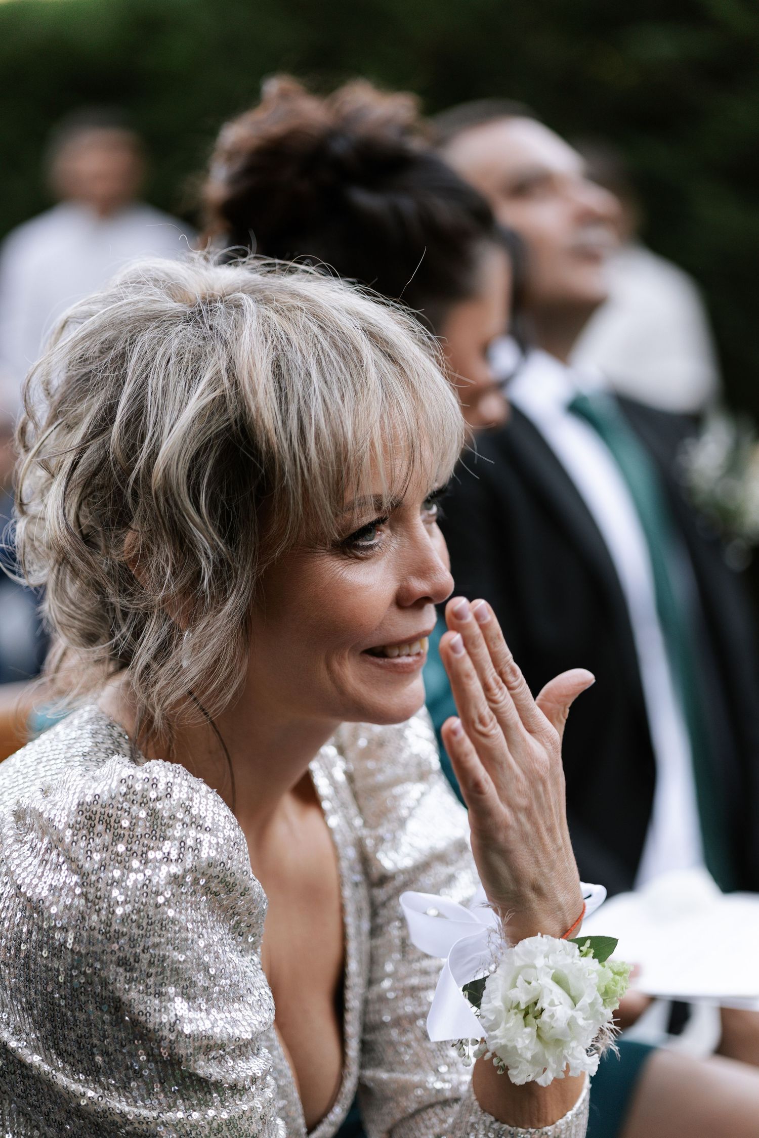 A guest at a wedding wearing a sparkly silver sequin dress and white floral wrist corsage reacts emotionally to the ceremony.