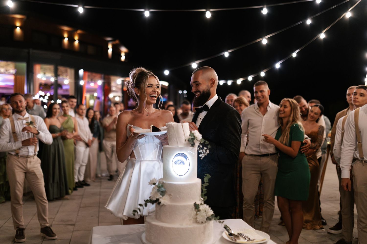A couple cuts their elegant white wedding cake under twinkling string lights at an outdoor evening reception.