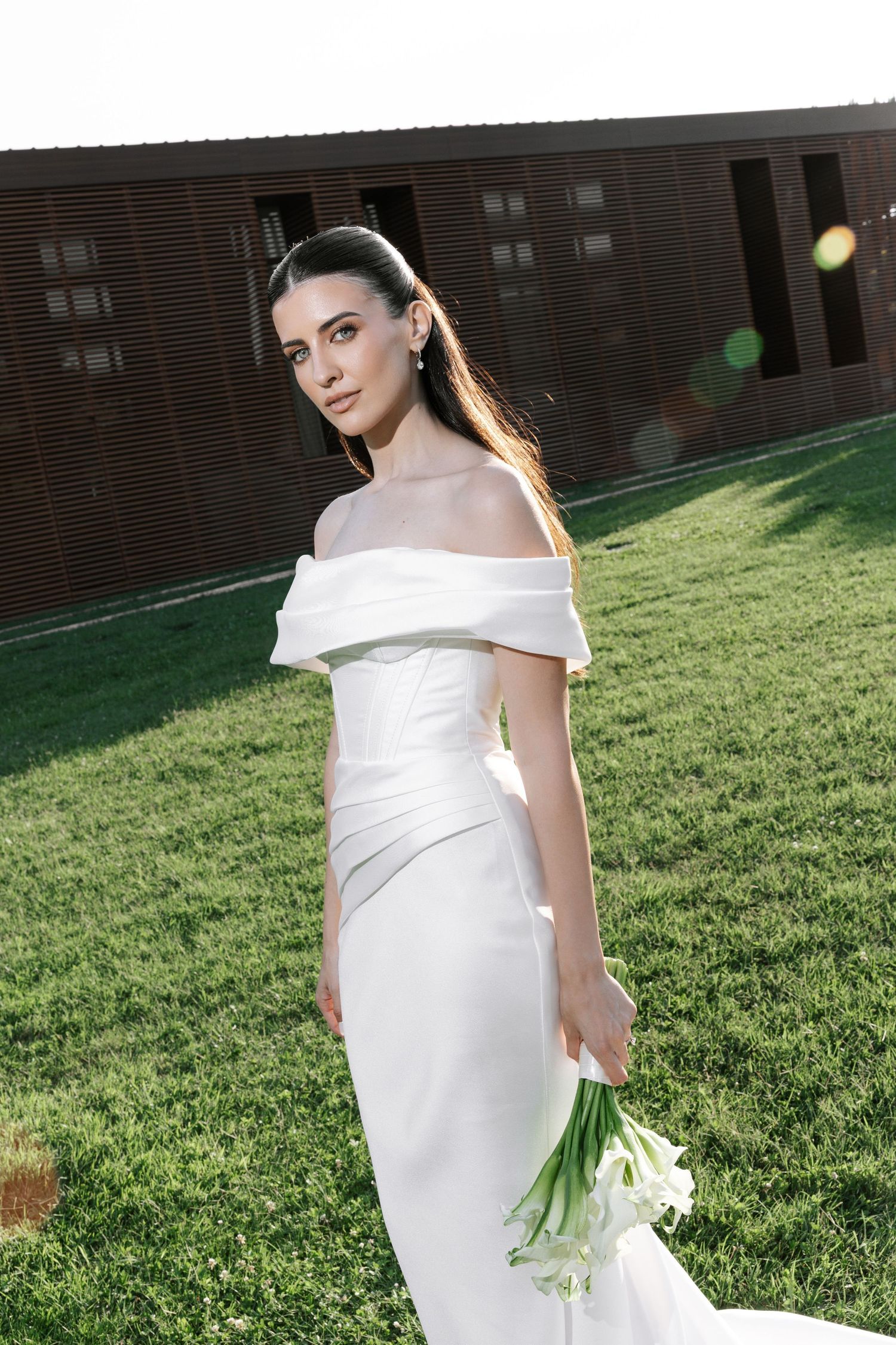 A bride in an elegant off-shoulder white wedding gown poses on a sunlit grassy lawn.