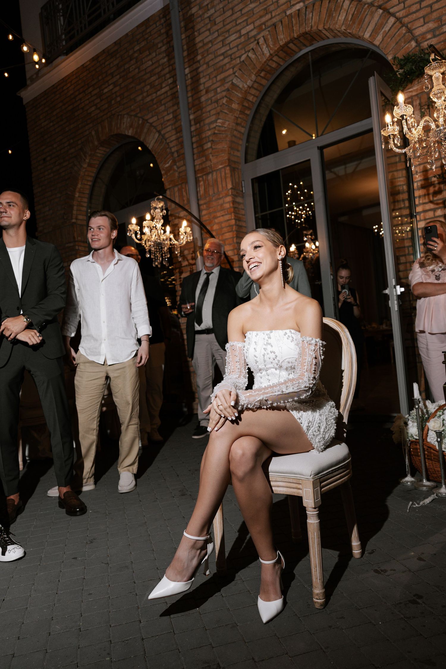 Wedding reception scene with guests gathered around bride in short white dress.