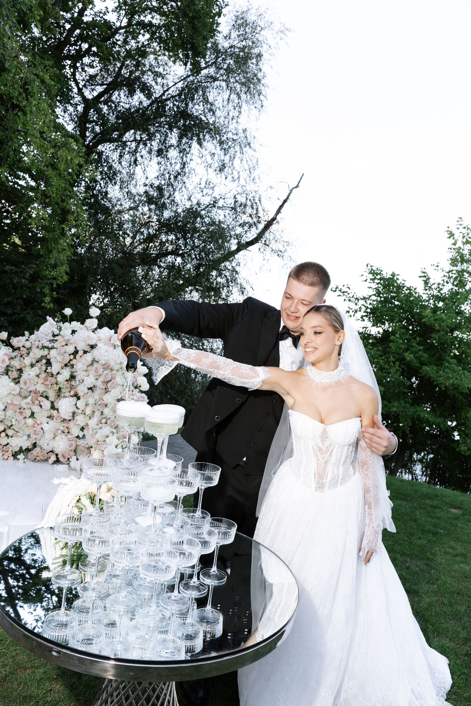 Wedding couple performing a champagne toast with a tower of glasses at an outdoor reception.