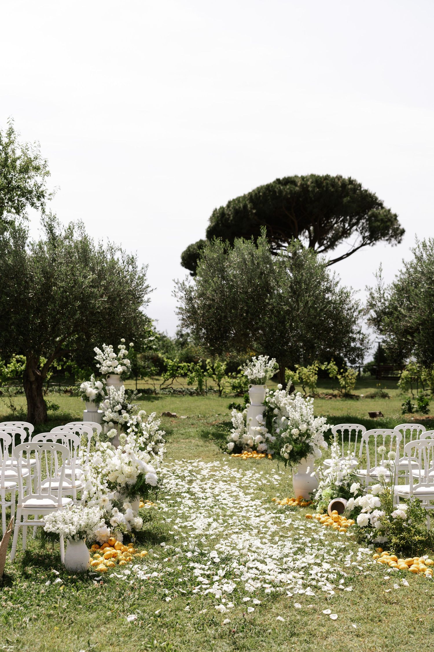 White chairs and floral arrangements line an outdoor garden path with scattered petals and rustic decor for a wedding ceremony.