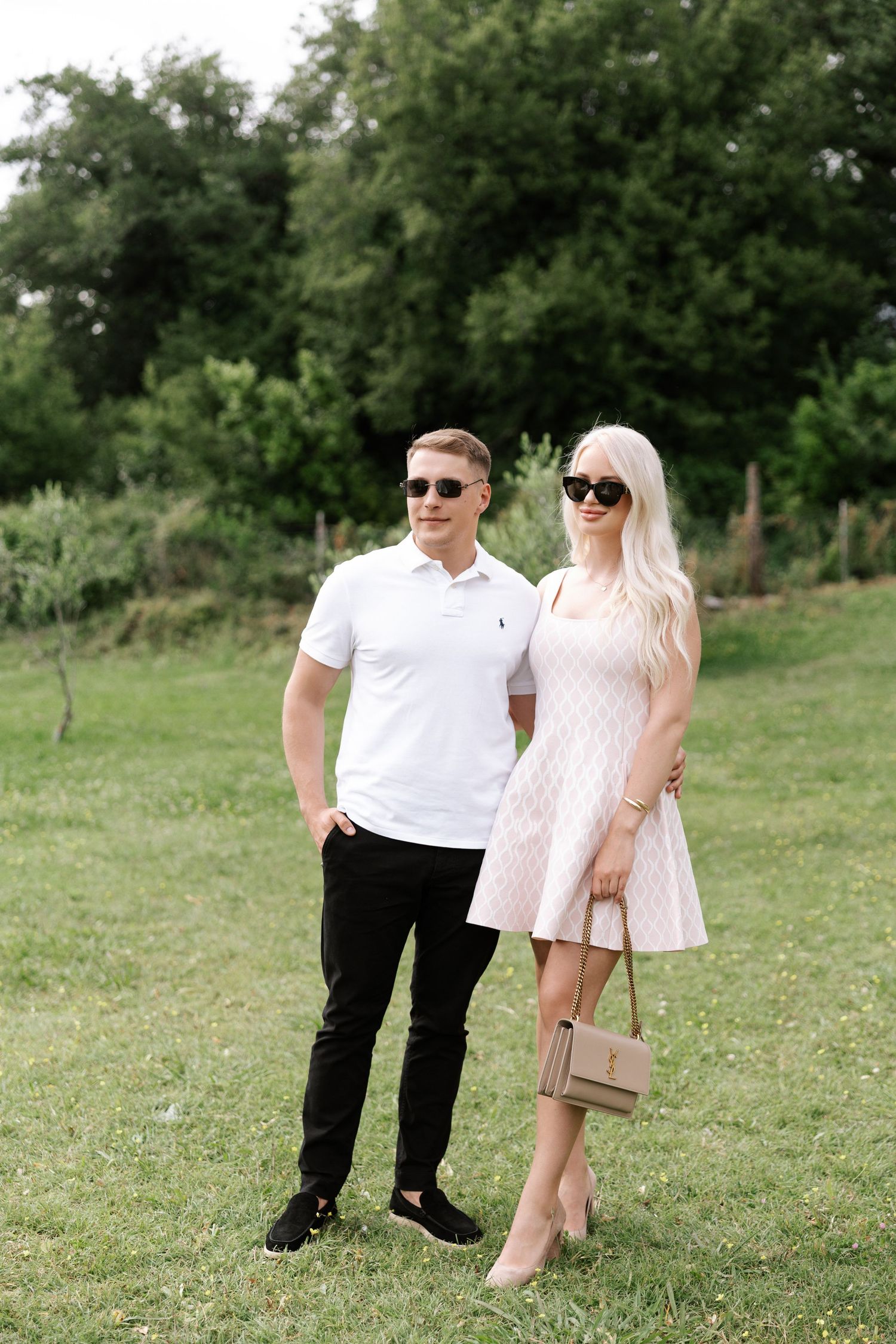 Two people in coordinated white and pink outfits stand together in a lush green field on a summer day.