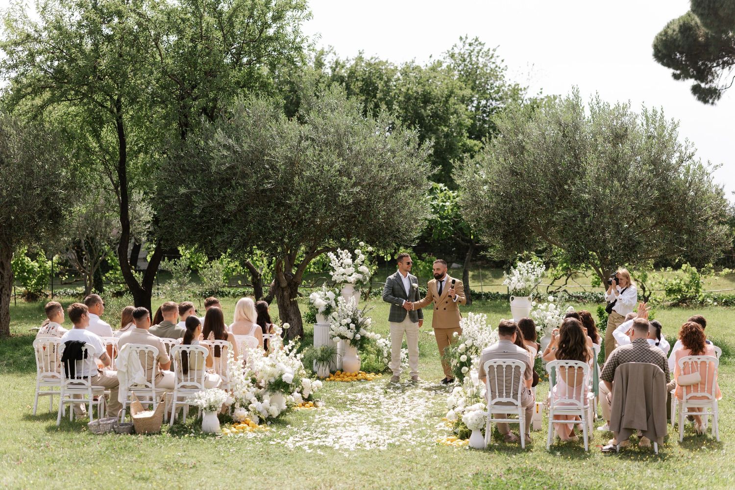 Intimate outdoor garden wedding ceremony under olive trees with white chairs and scattered flower petals on the grass.