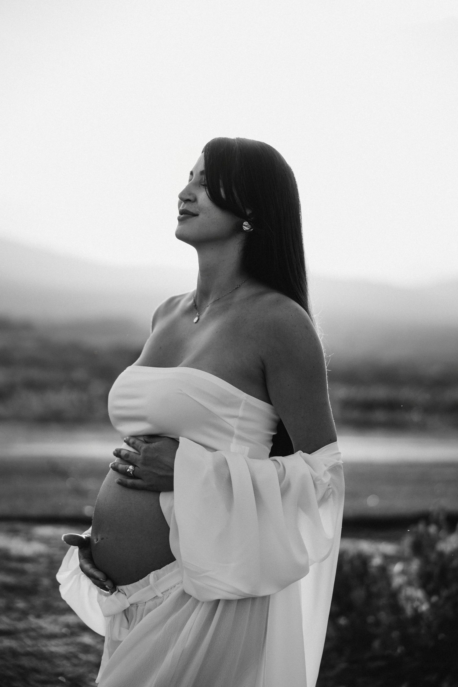 Expecting mother in flowing white dress poses against scenic mountain backdrop in black and white photograph.