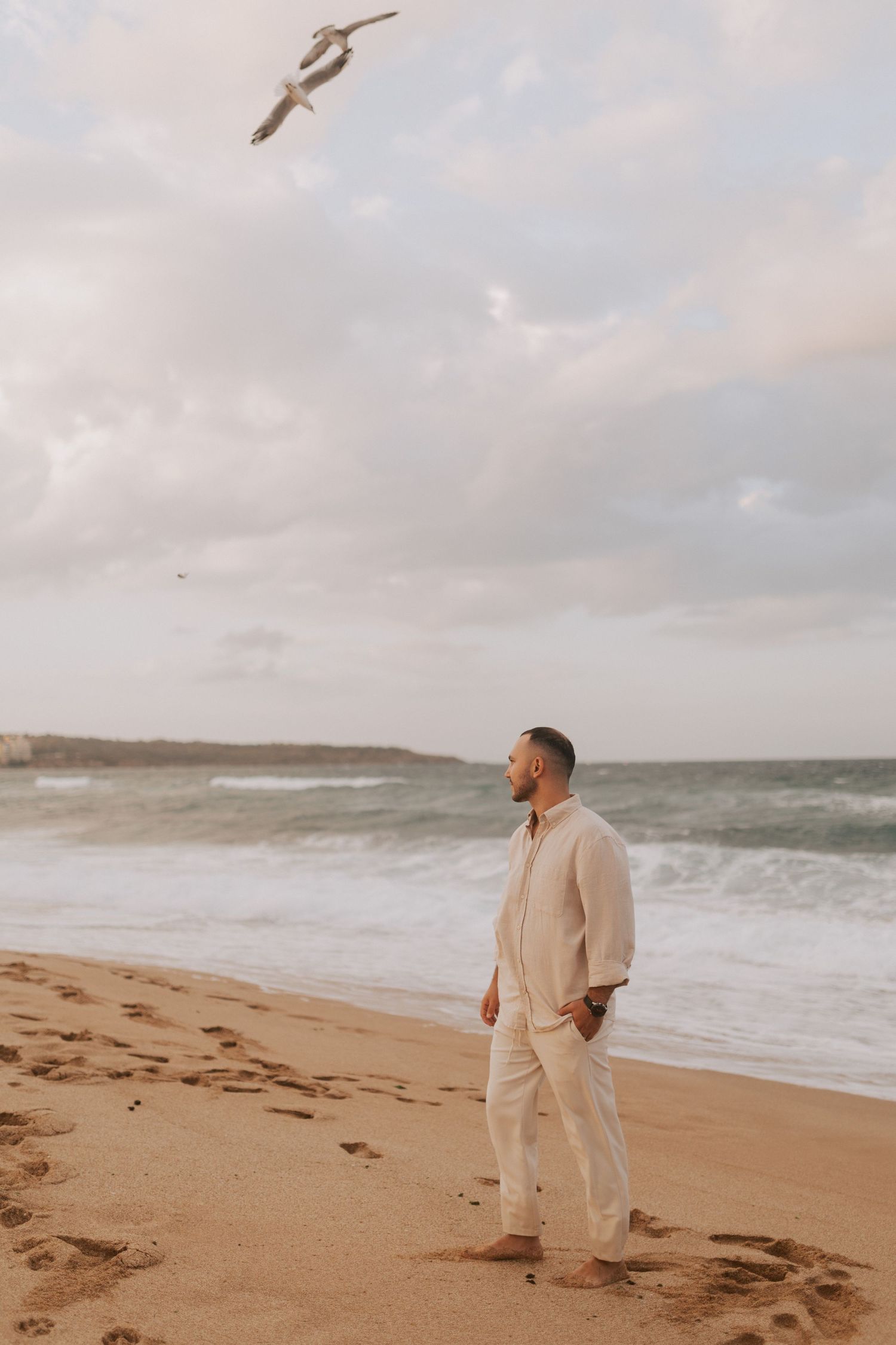A person in light clothing watches a seabird soar above a dramatic beach landscape.