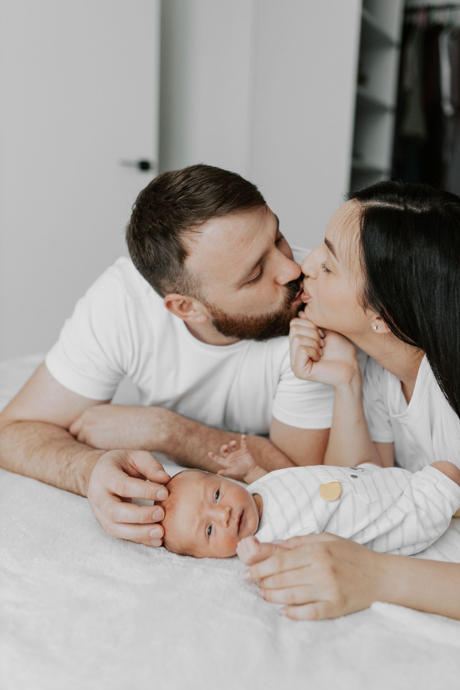An intimate family portrait on white bedding with a sleeping baby between loving parents.