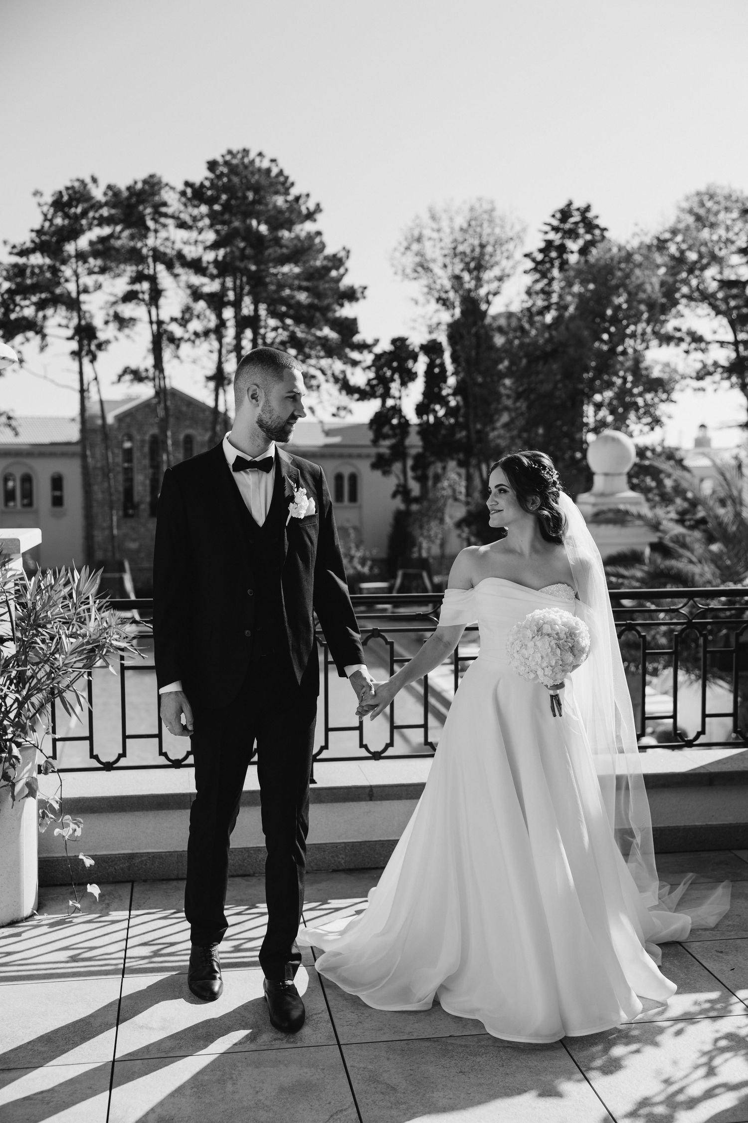 Black and white portrait of newlyweds on balcony with trees and buildings in background.