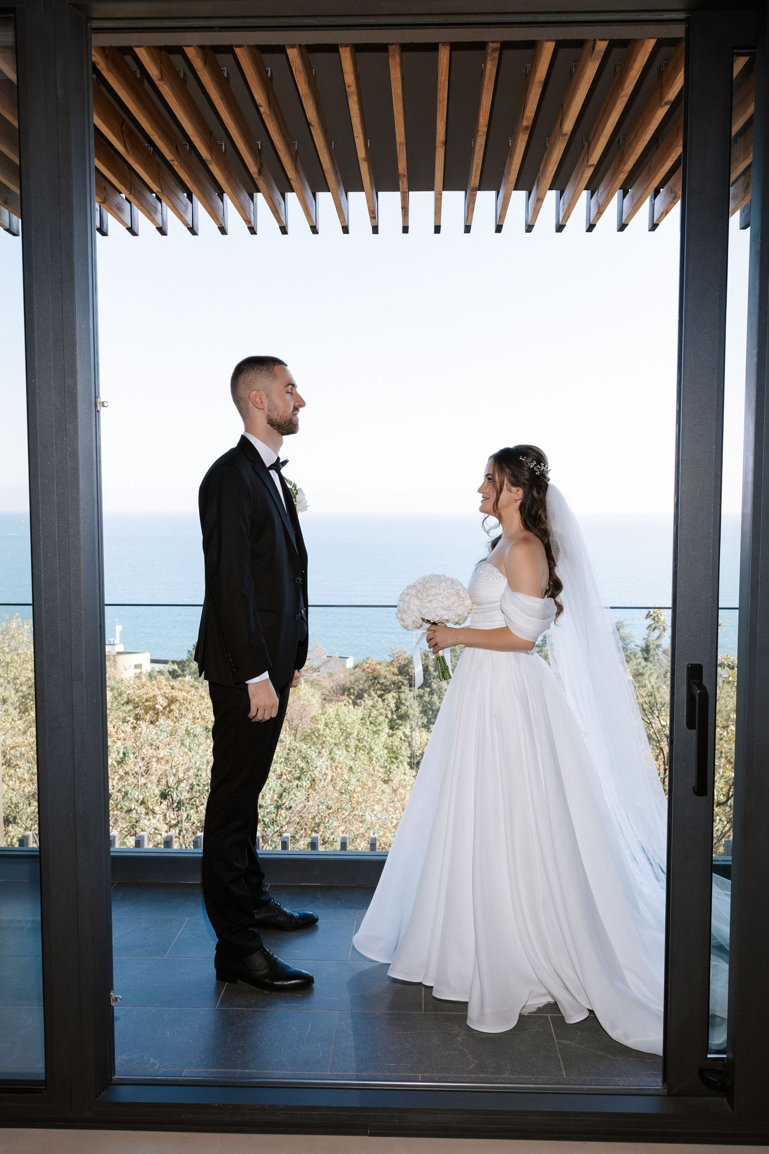 Couple stands under wooden pergola overlooking ocean view on their wedding day.
