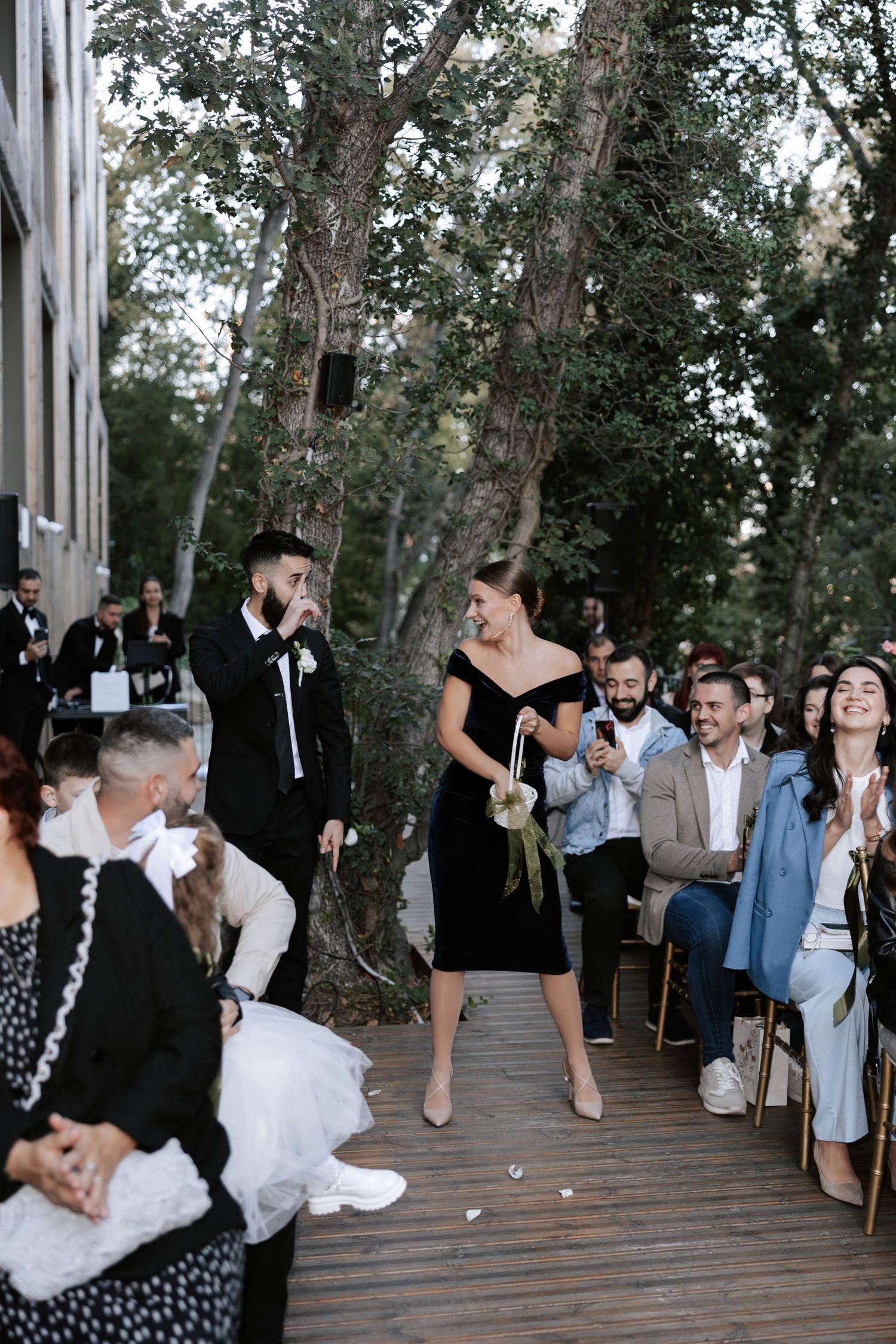 Guests observe a wedding celebration on a wooden deck surrounded by trees.