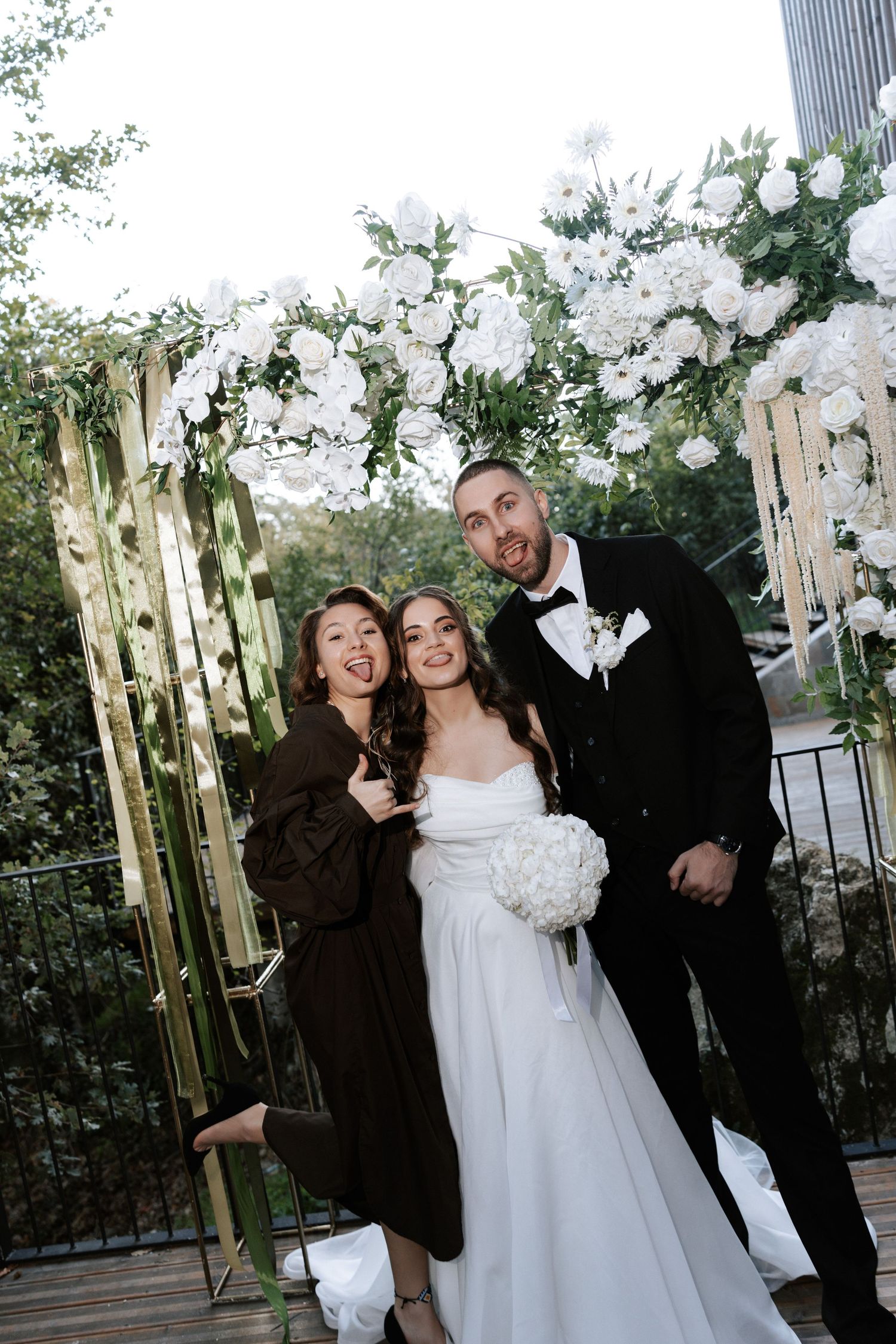 Wedding party poses together under decorative white floral arch on wooden deck.