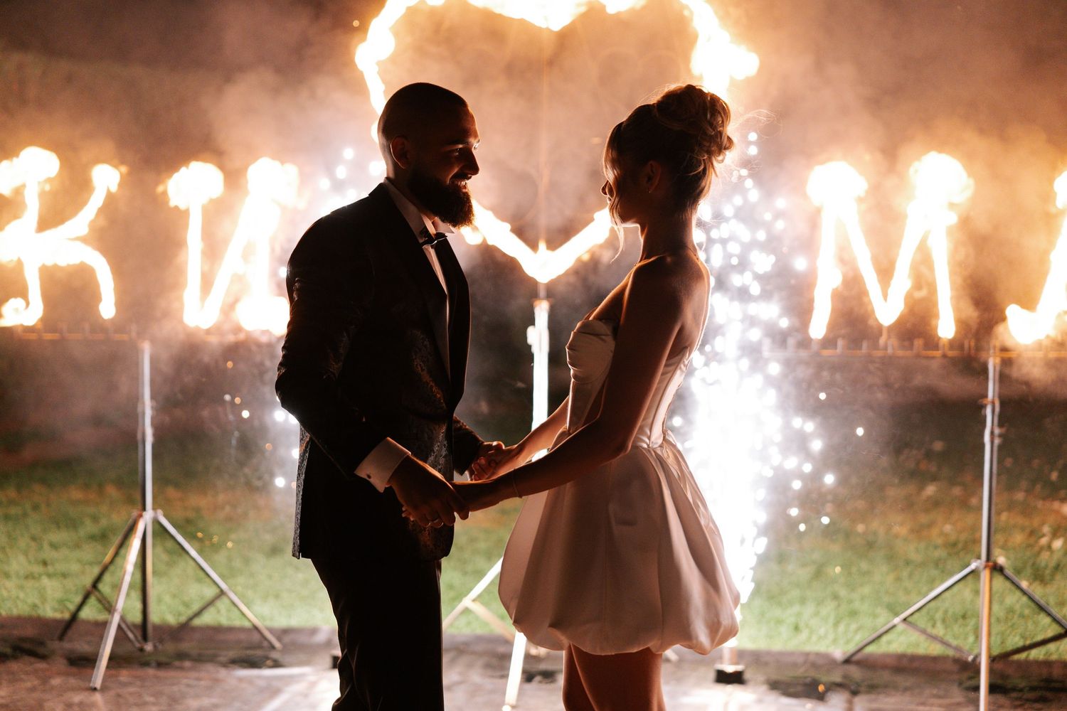 A couple stands in silhouette against glowing letters and sparklers during a romantic nighttime celebration.