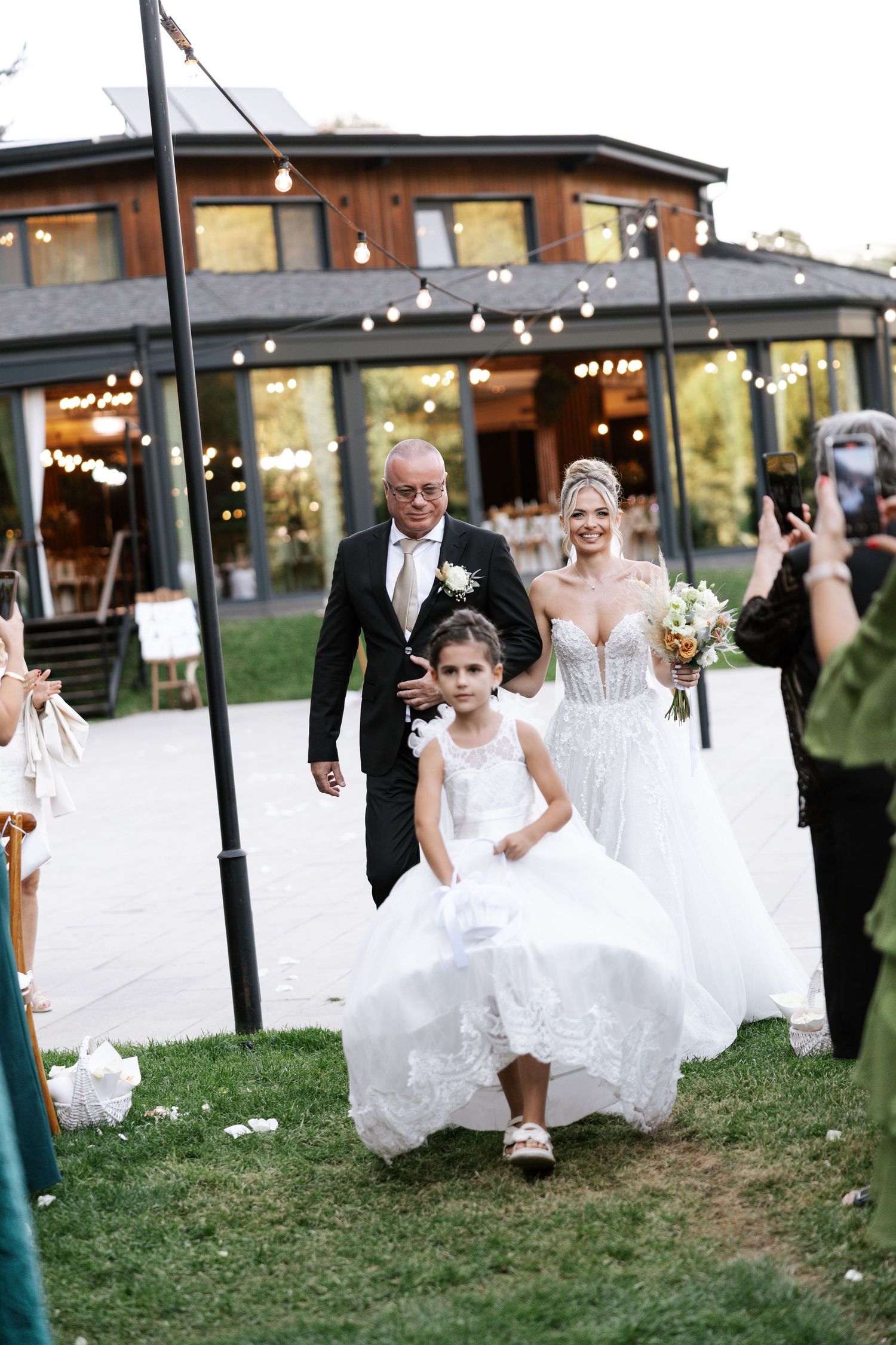 A group of brides in white wedding dresses run together across a grassy lawn at an outdoor venue.