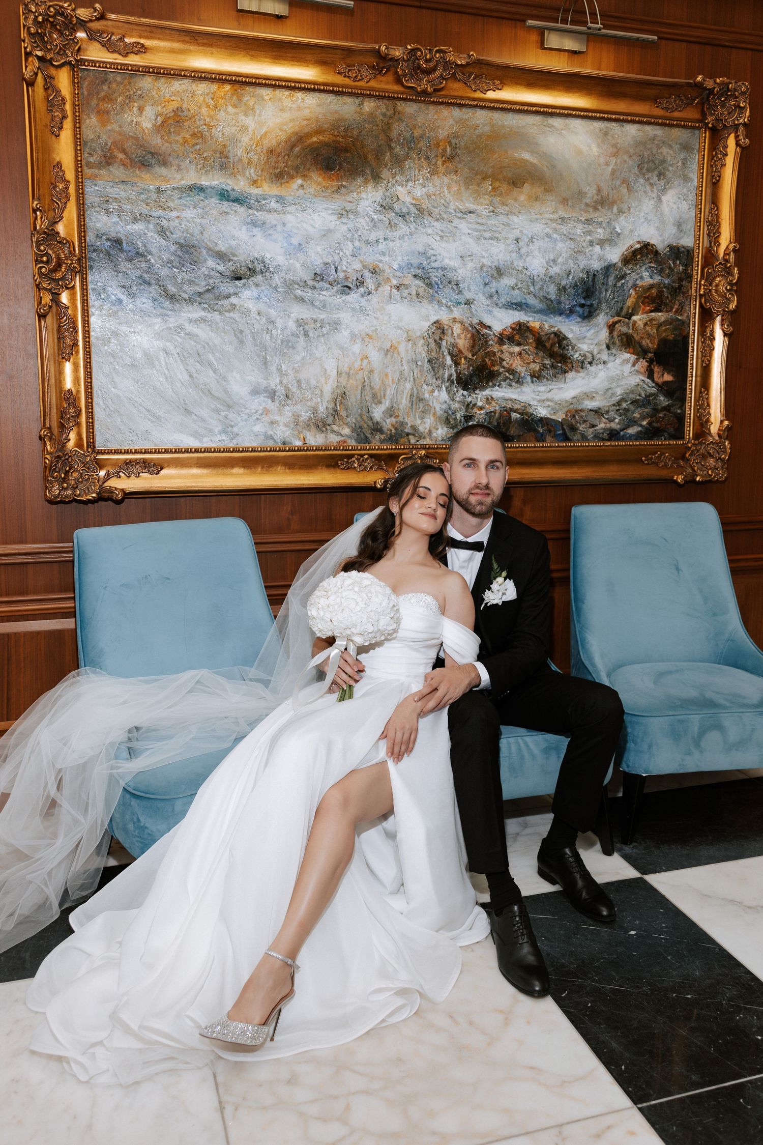 Elegant wedding portrait with couple seated beneath large ocean artwork in gold frame.