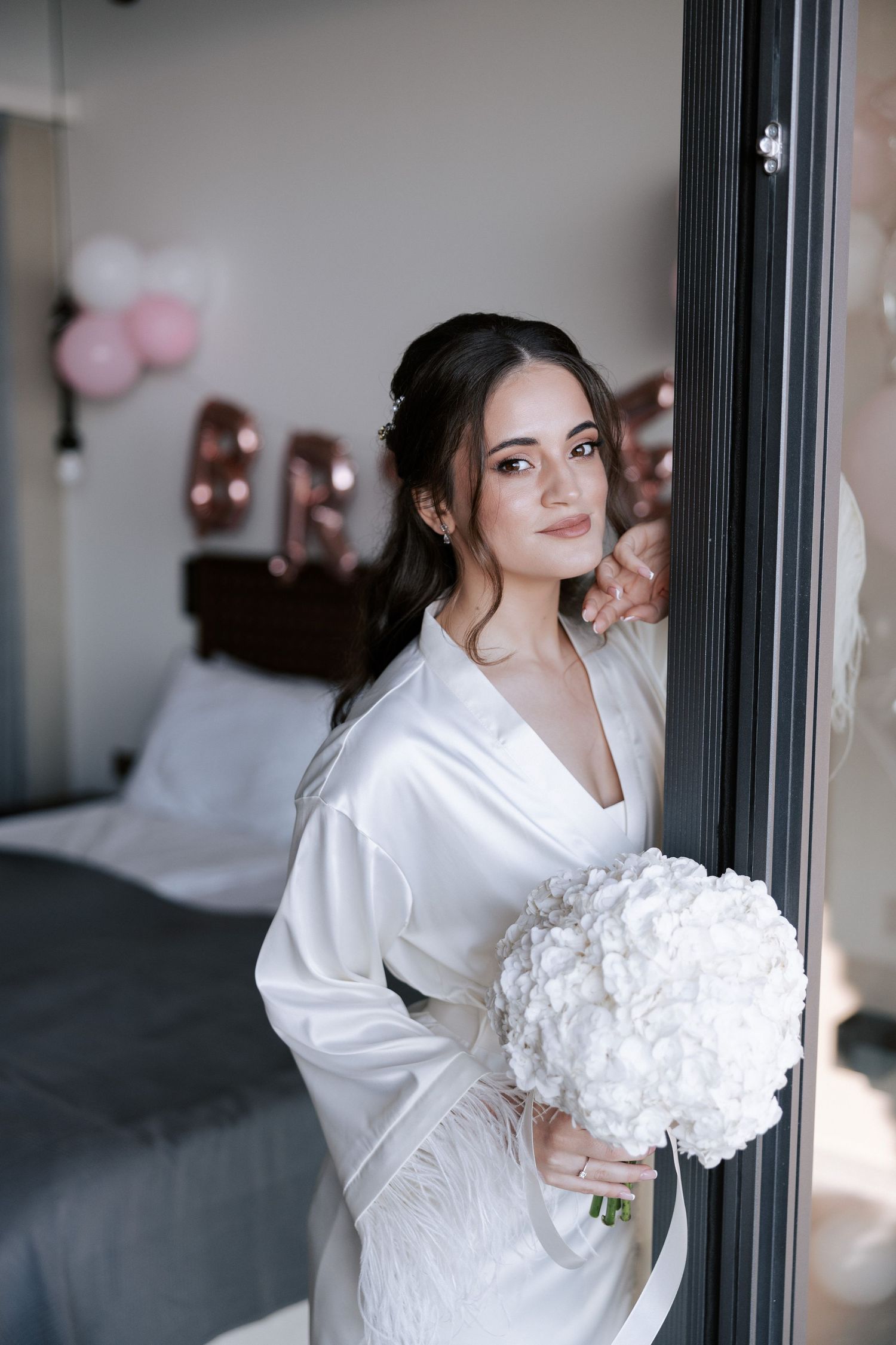 Person in white silk robe holding white bouquet against bedroom interior.