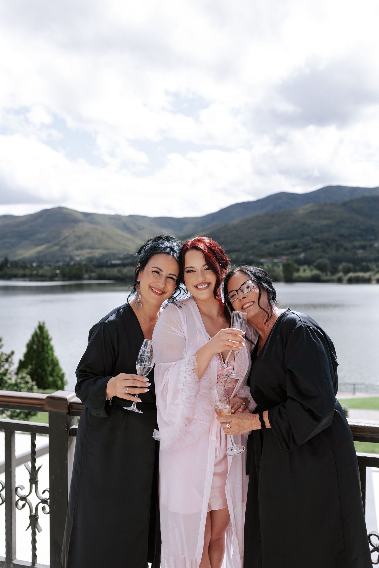 Three women in robes share champagne on balcony with mountain lake view.Wedding photographer Bulgaria