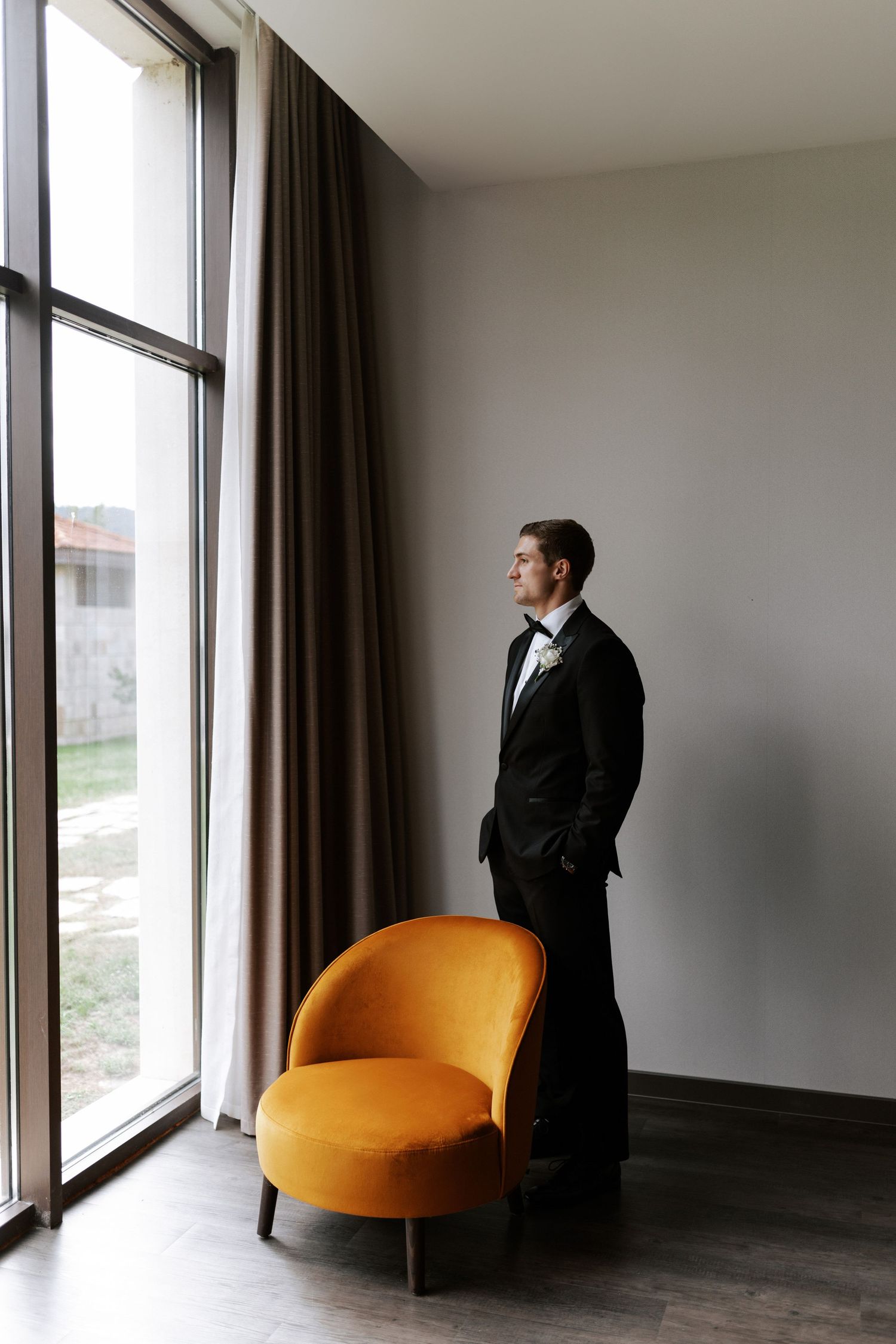 A person in formal attire stands next to an orange velvet chair by a floor-to-ceiling window.