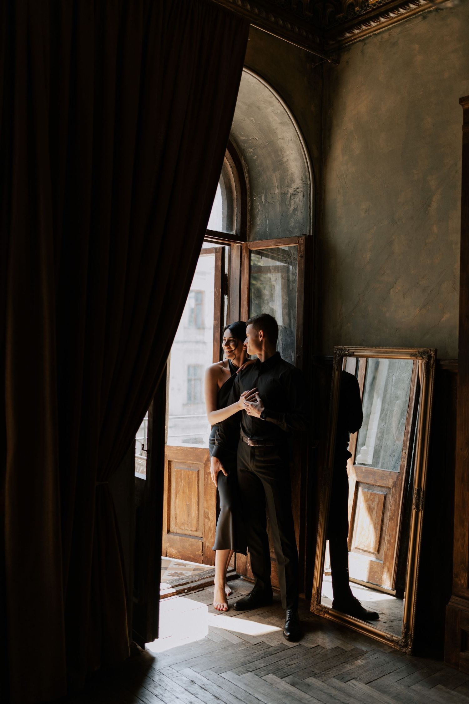 A couple embraces by an arched window in a dimly lit vintage room with dark curtains and wooden details.