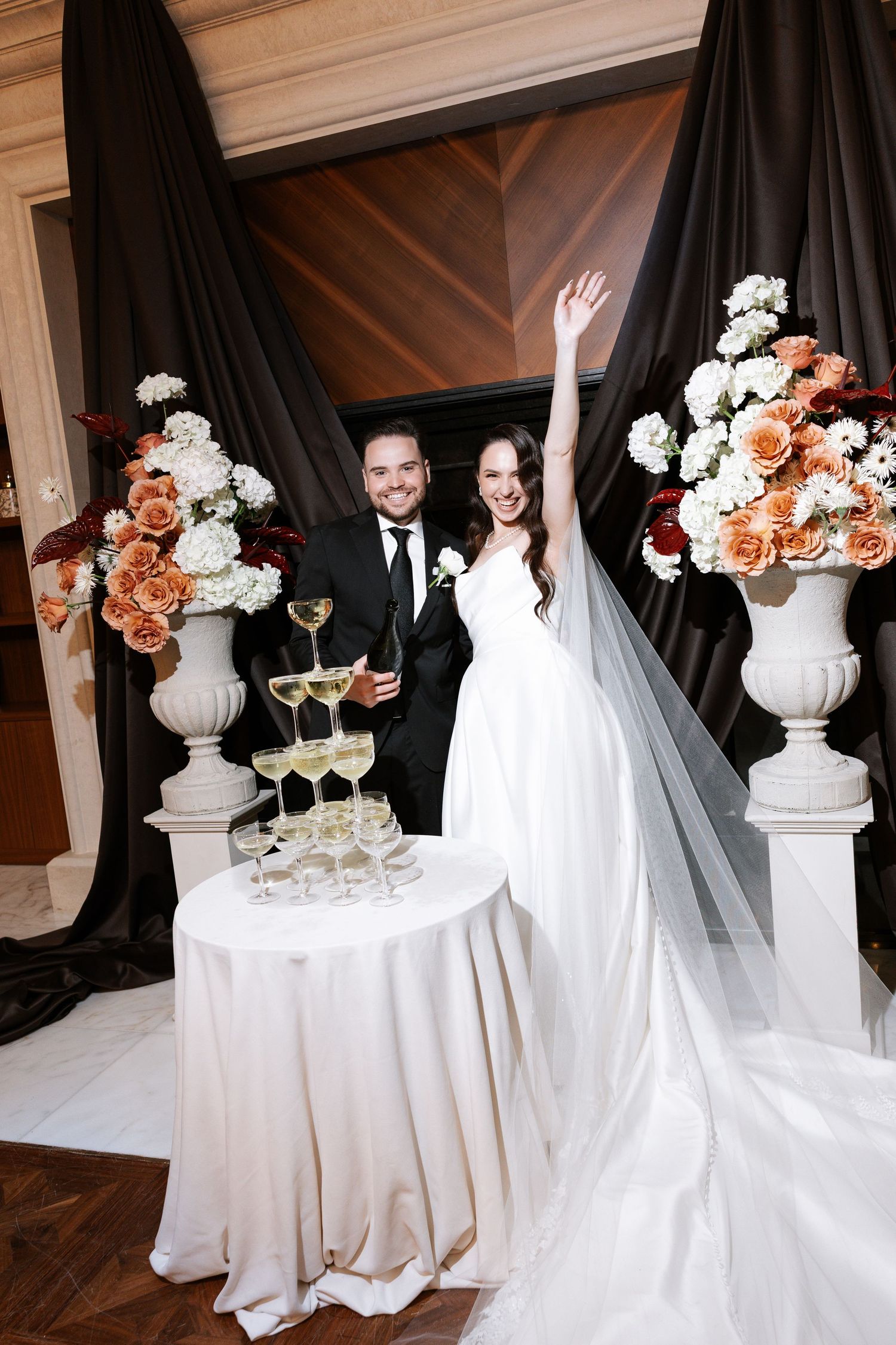 Elegant wedding couple poses in formal attire with white and peach floral arrangements and champagne glasses.