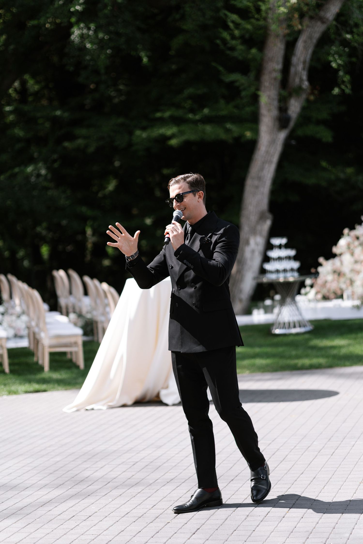 Person in black suit gesturing while speaking at outdoor wedding venue.