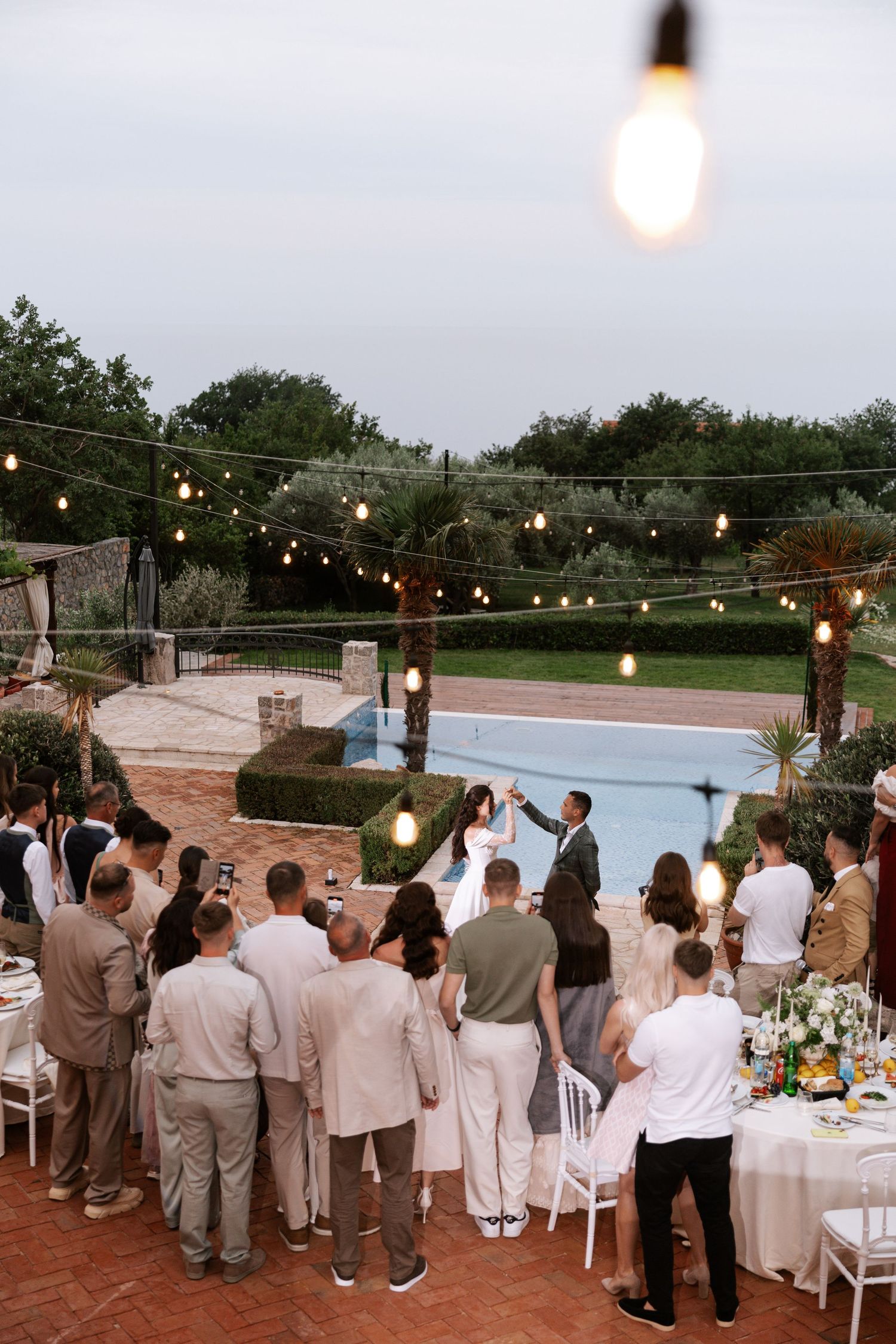 Wedding guests gather outdoors at dusk under string lights for a celebration on a patio overlooking a scenic view.