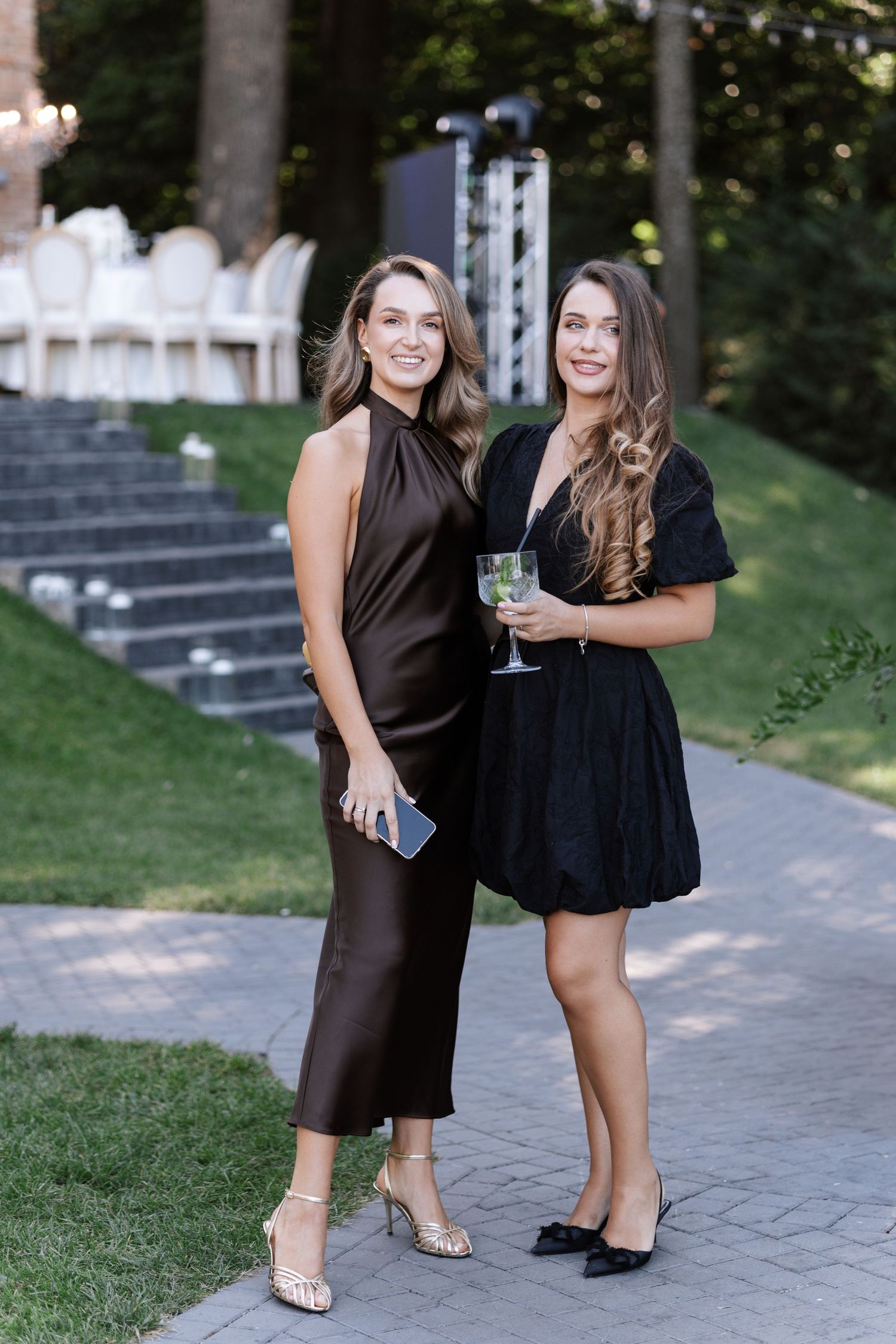 Two guests in formal attire posing together on a stone pathway at an outdoor event.