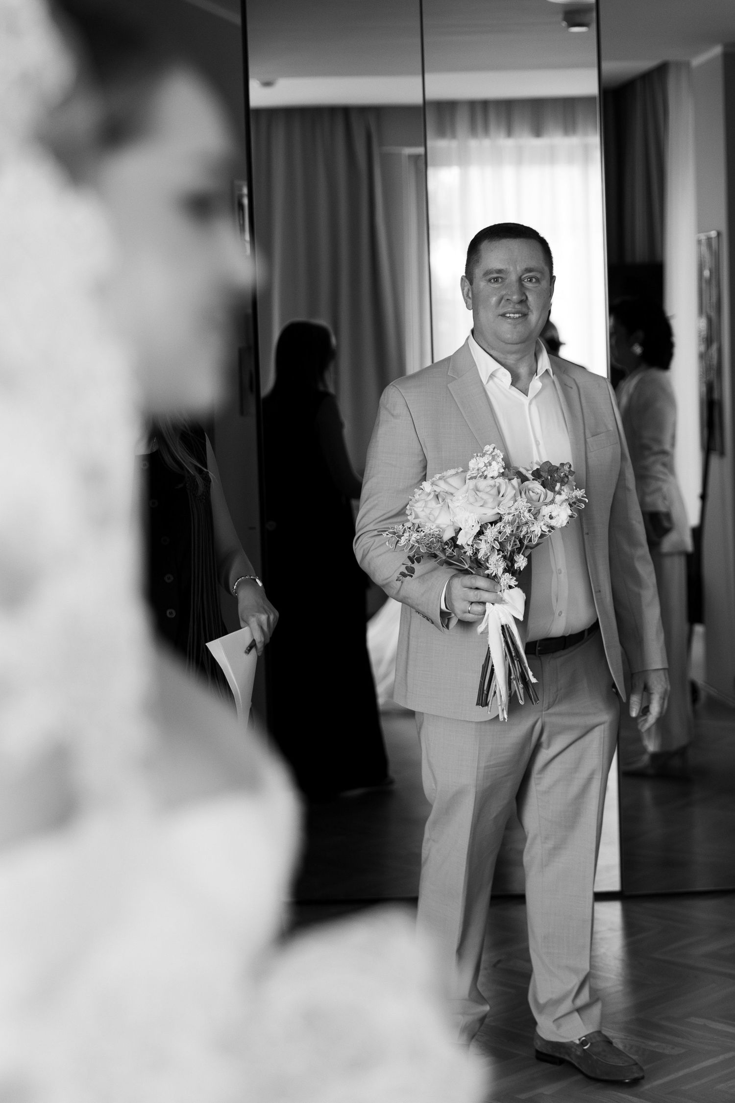 A person in a gray suit carries a delicate bouquet of white flowers down a hallway.