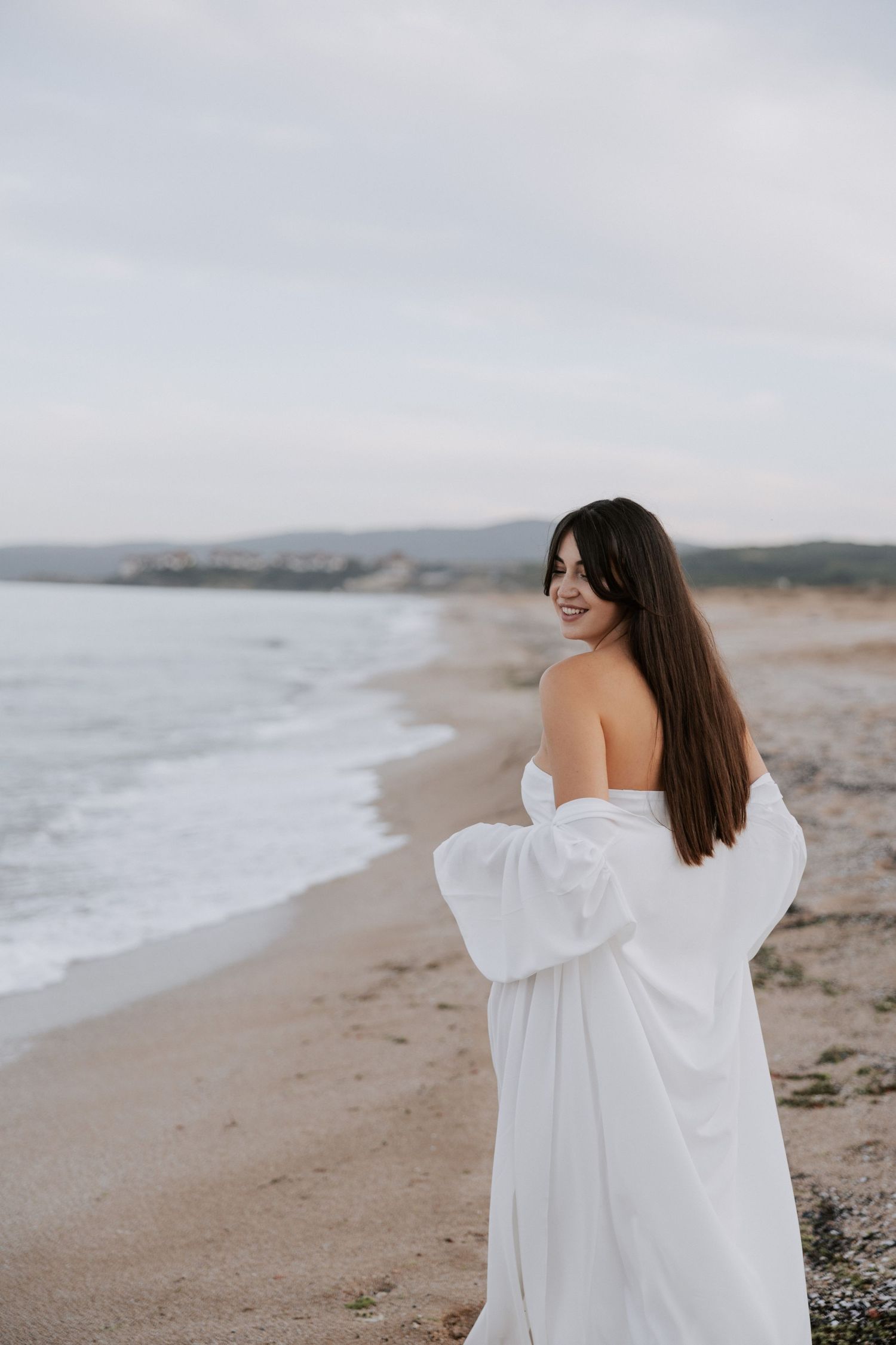 A pregnant woman in a flowing white dress stands on a beach looking over her shoulder.